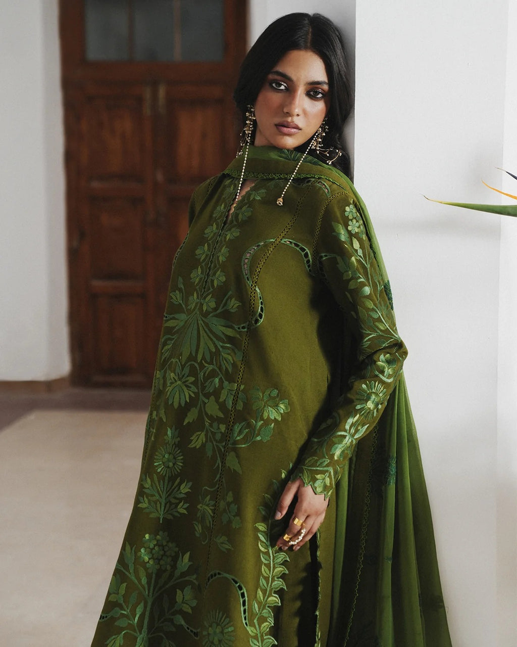 A woman models a green embroidered kurta set with a matching sheer dupatta in an indoor setting with a wooden door in the background.