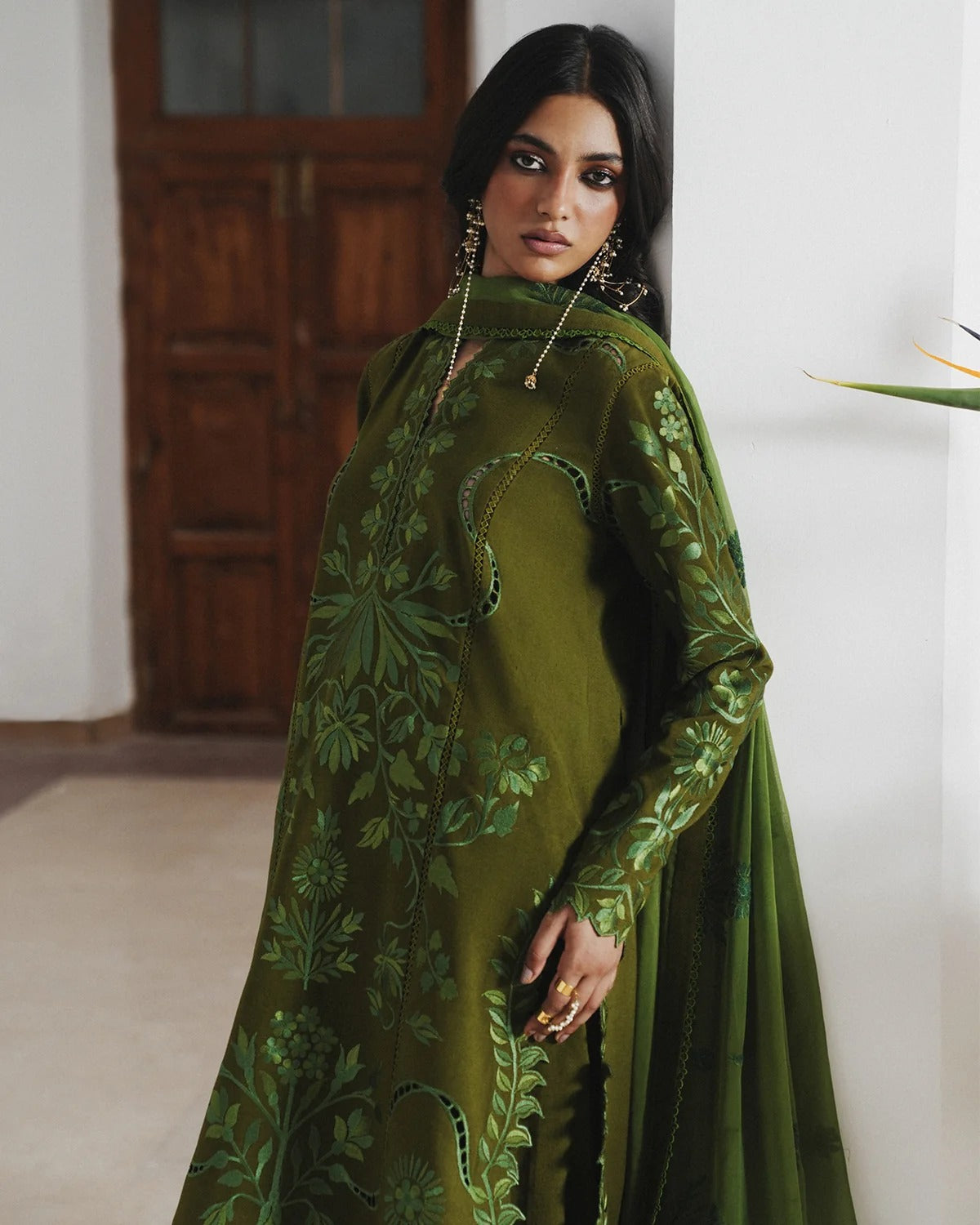 A woman models a green embroidered kurta set with a matching sheer dupatta in an indoor setting with a wooden door in the background.