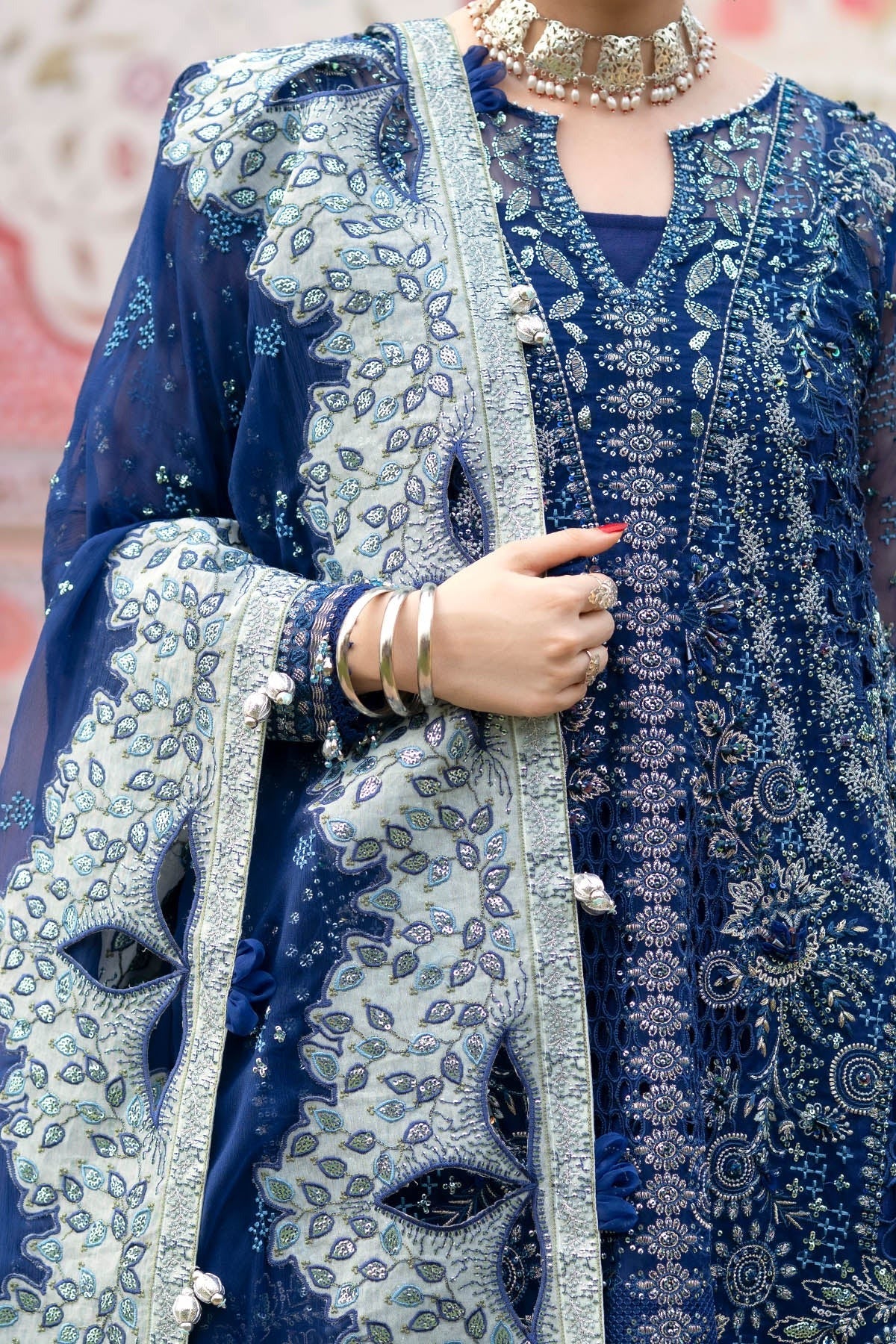 A close-up of a person wearing a richly embroidered navy blue traditional outfit with a matching sheer dupatta featuring intricate silver and white floral patterns, accessorized with a gold necklace and bangles.