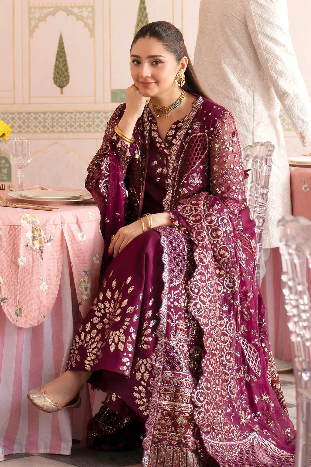 A woman models a traditional deep magenta three-piece ethnic outfit with intricate gold embroidery, seated in an elegant indoor setting with a pink patterned tablecloth.