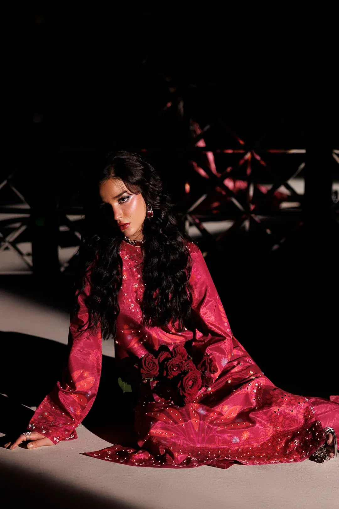 A woman wearing a long-sleeved, floor-length magenta pink embroidered dress with sequins and rose motifs, kneeling on a light-colored floor in a dark, moody setting.