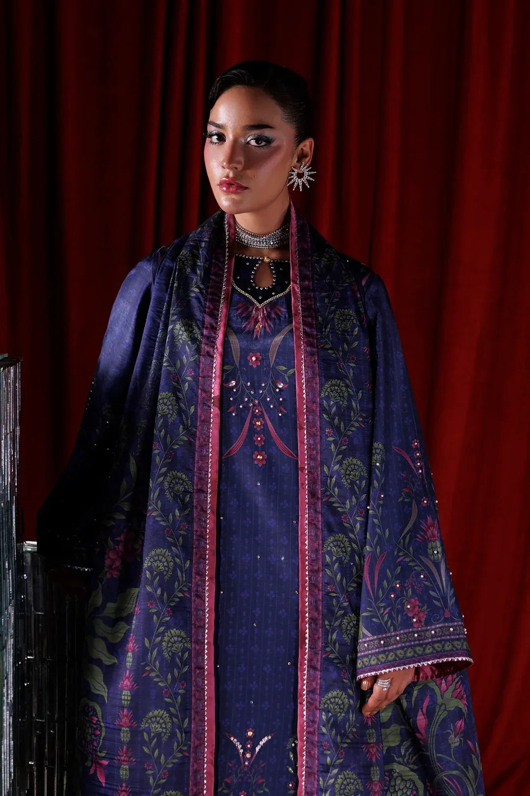 A model wearing a traditional South Asian outfit featuring a navy blue kurta with intricate pink and green floral embroidery, paired with a matching embroidered shawl, set against a dark red curtain background.
