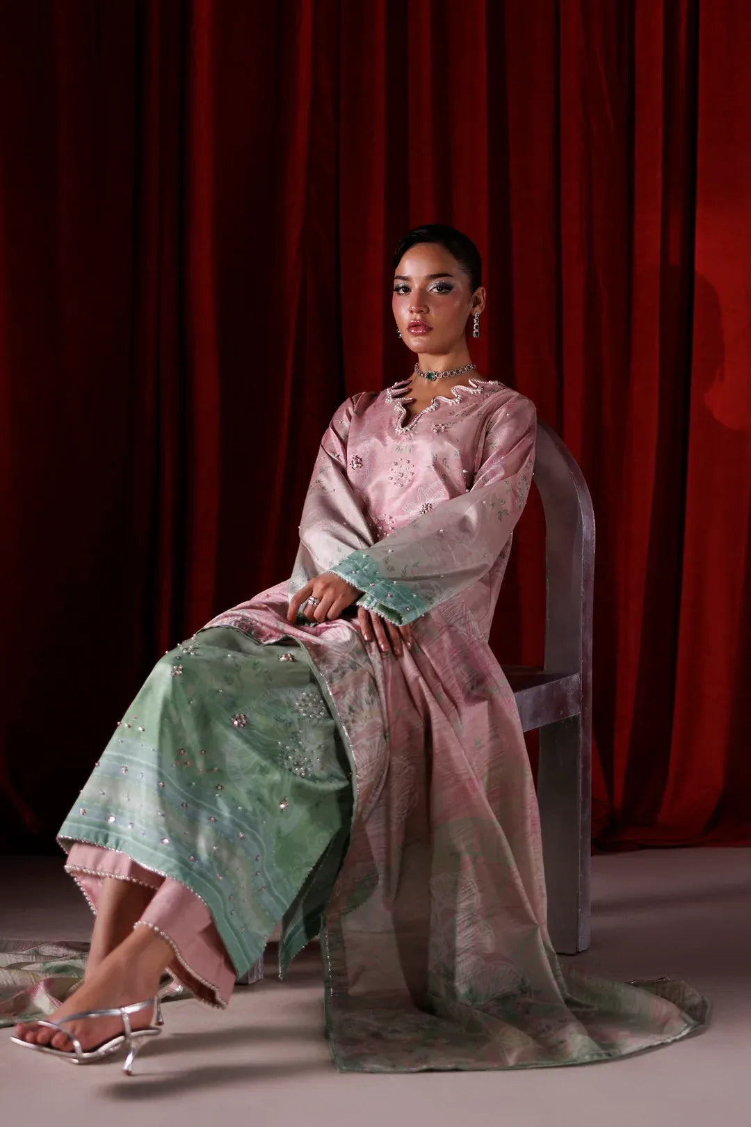A woman seated on a chair in front of a red curtain, wearing a traditional outfit with a light pink embroidered top and a flowing mint green skirt with metallic embellishments.