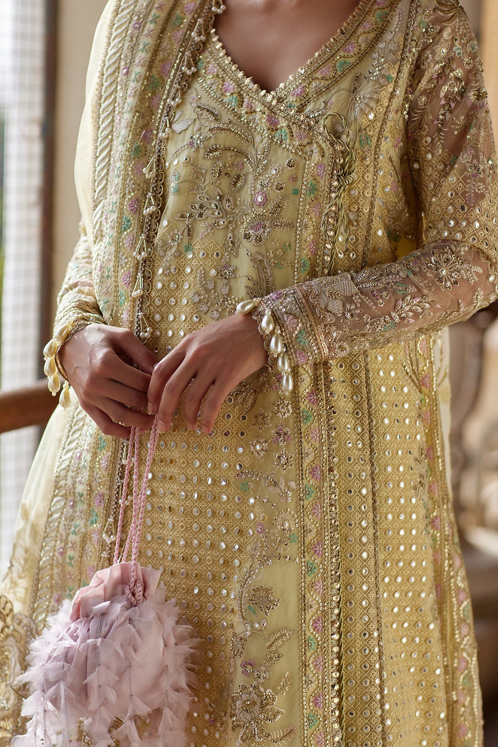A close-up of a person wearing an ornate beige and gold embroidered traditional outfit with mirror work and bead detailing, holding a small pink feathered handbag.