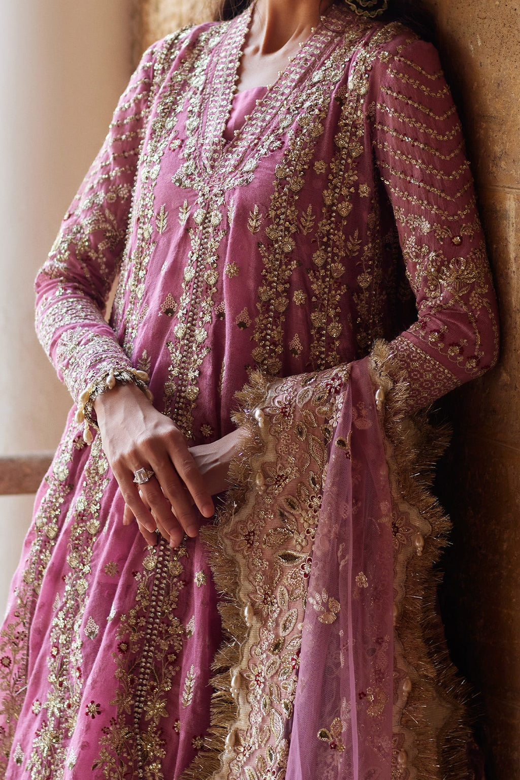 A close-up of a person wearing a pink traditional outfit with extensive gold embroidery and a matching sheer dupatta draped over one arm, set against a warm neutral background.