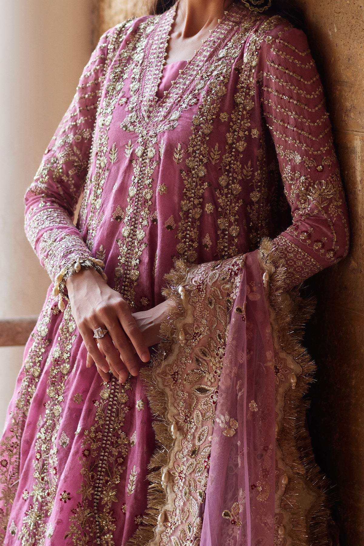 A close-up of a person wearing a pink traditional outfit with extensive gold embroidery and a matching sheer dupatta draped over one arm, set against a warm neutral background.