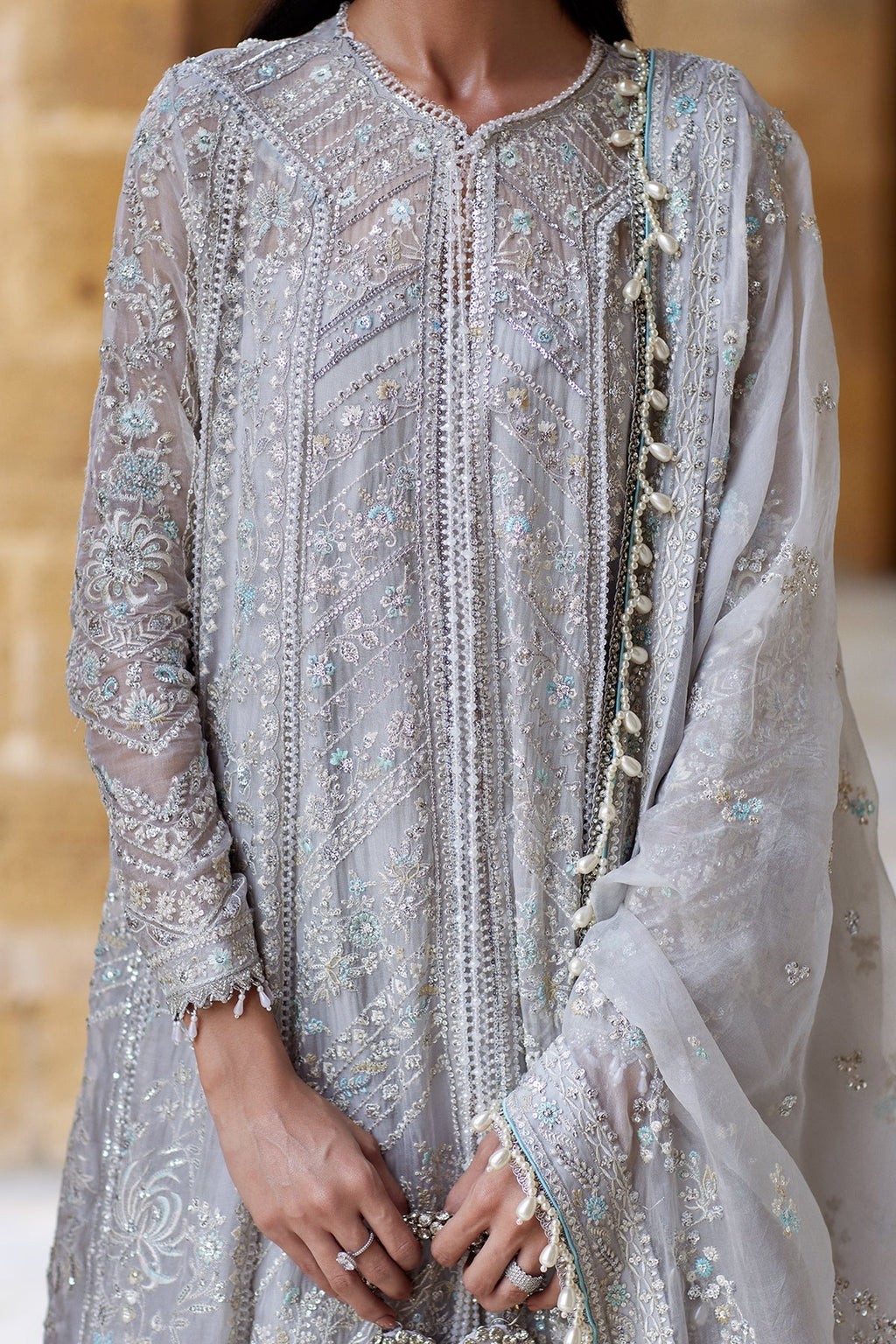 A close-up of a person wearing a heavily embroidered light gray traditional outfit with intricate silver and blue beadwork, sheer sleeves, and a matching dupatta draped over one shoulder.