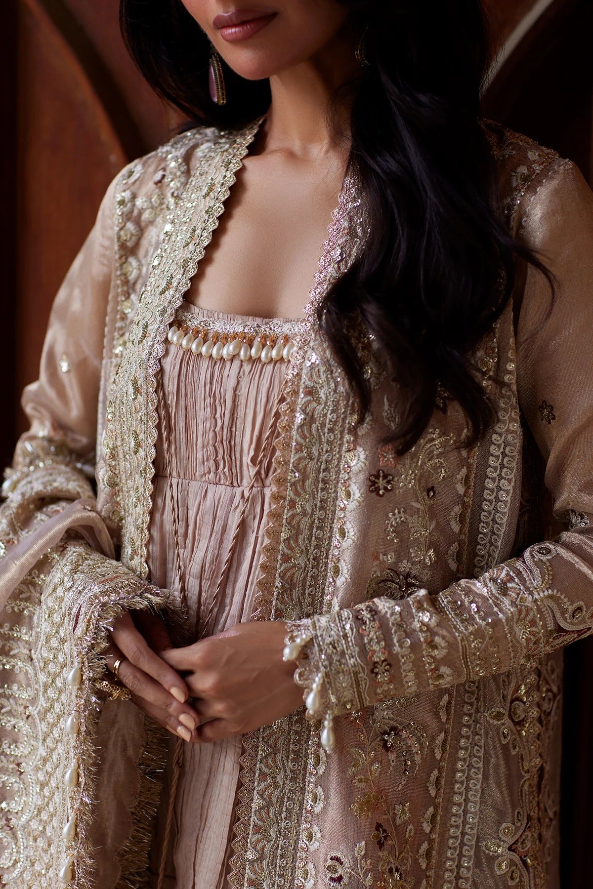A close-up of a woman wearing an ornate beige and gold traditional outfit with intricate embroidery, a matching inner dress, and a draped shawl.