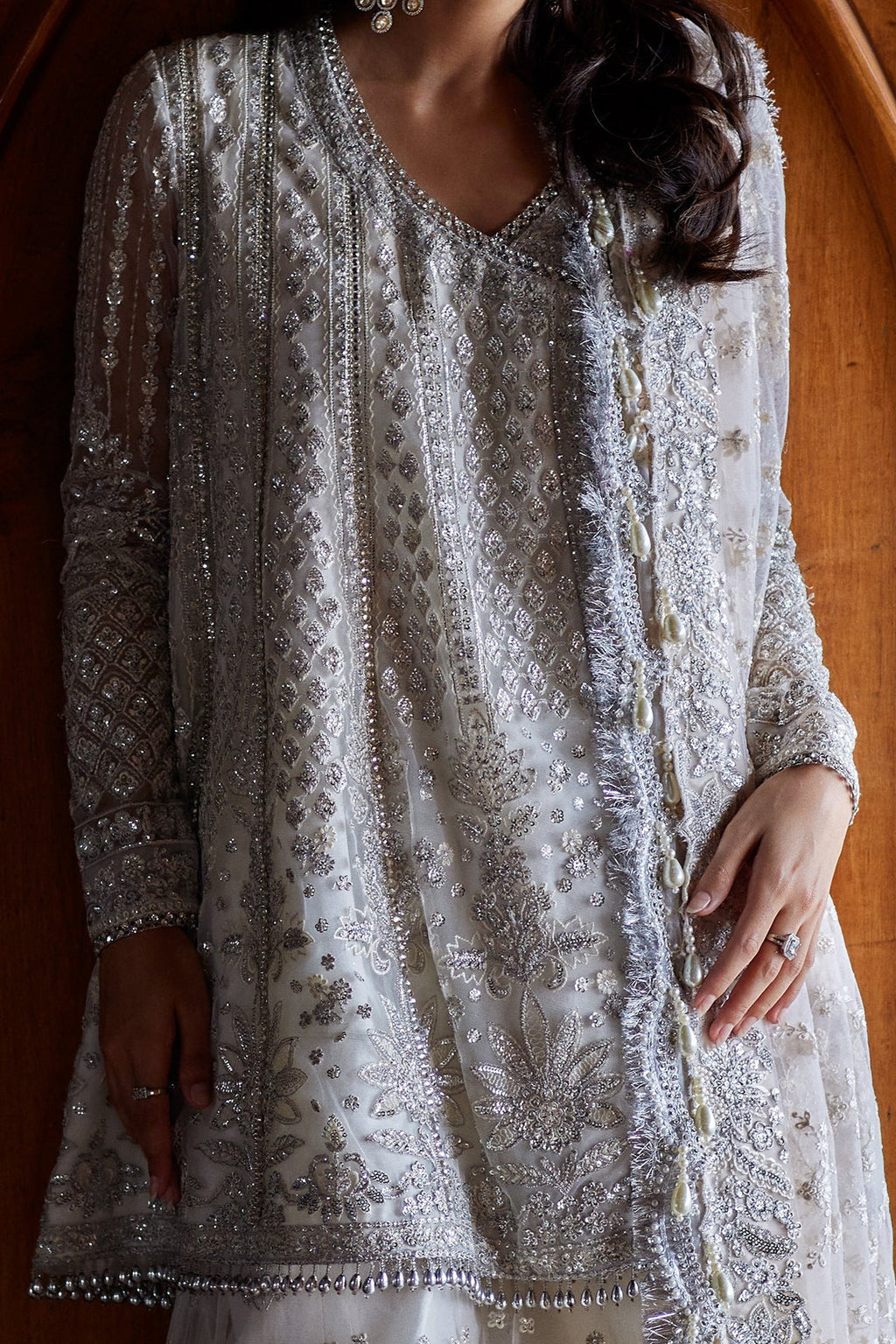 A close-up of a woman wearing a heavily embellished silver and white traditional tunic with intricate beadwork and embroidery, set against a warm wooden background.