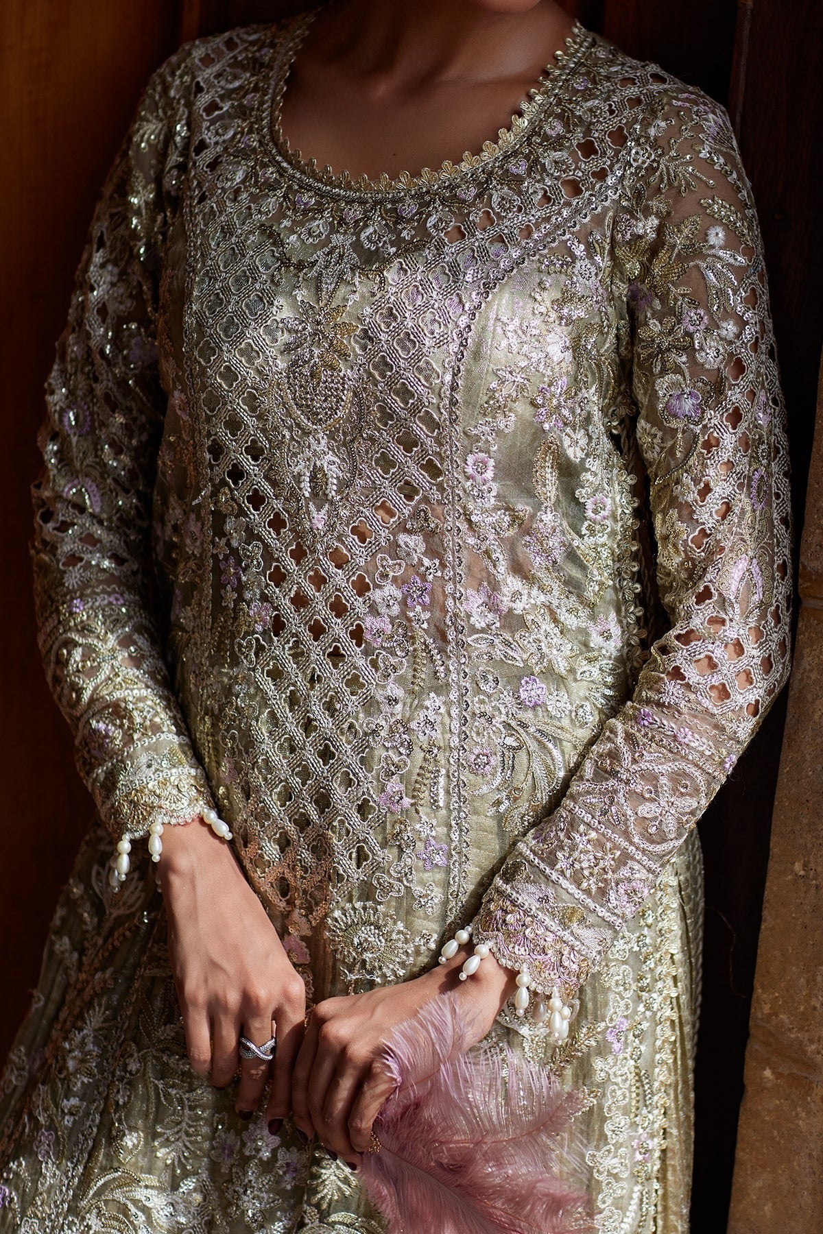 A close-up of a person wearing an ornate, heavily embroidered traditional outfit in shimmering metallic gold and silver with intricate floral lace detailing. The person is holding a pink feathered clutch against a dark wooden background.