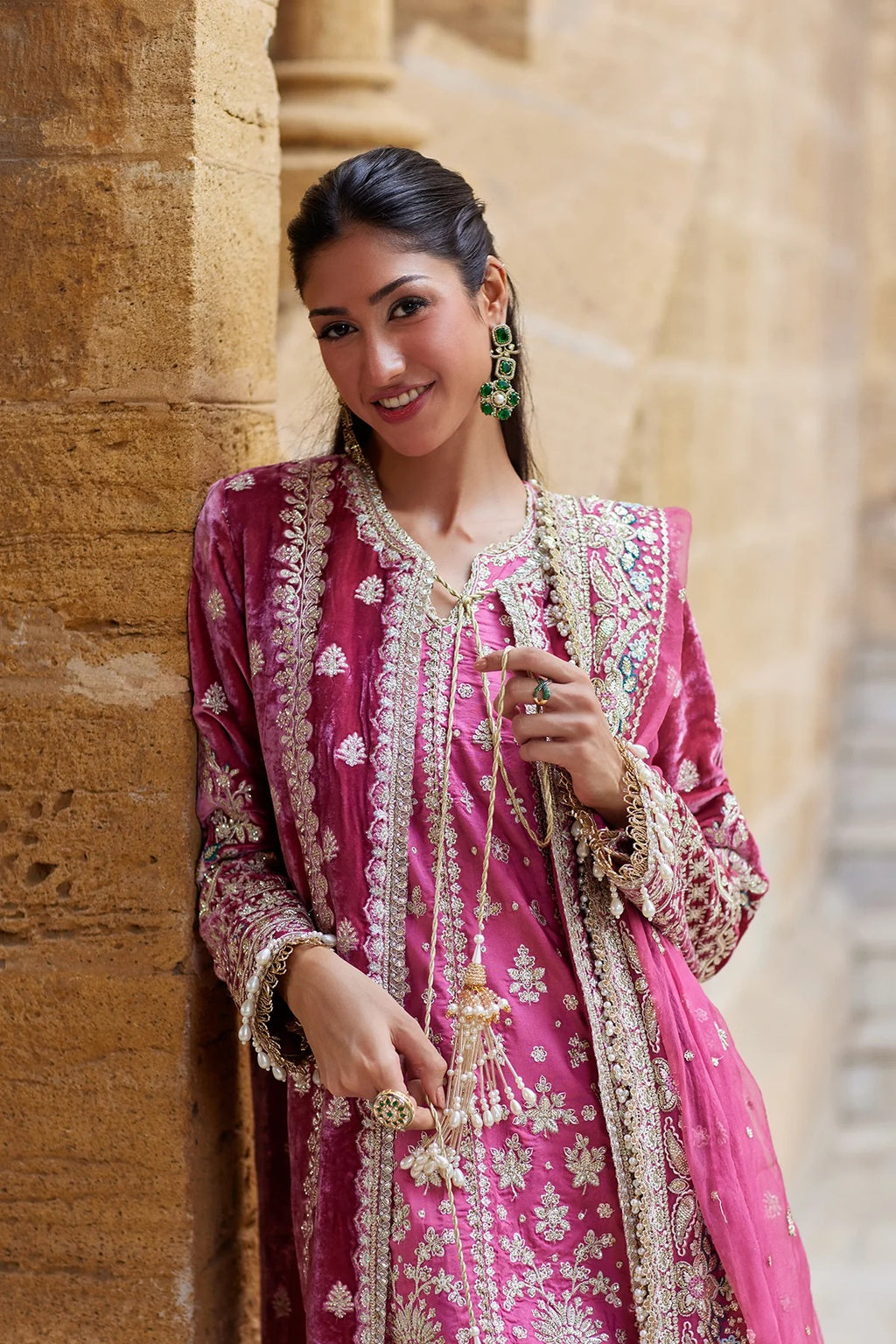 A woman wearing a vibrant pink traditional embroidered outfit with intricate gold detailing, standing against a beige stone wall.
