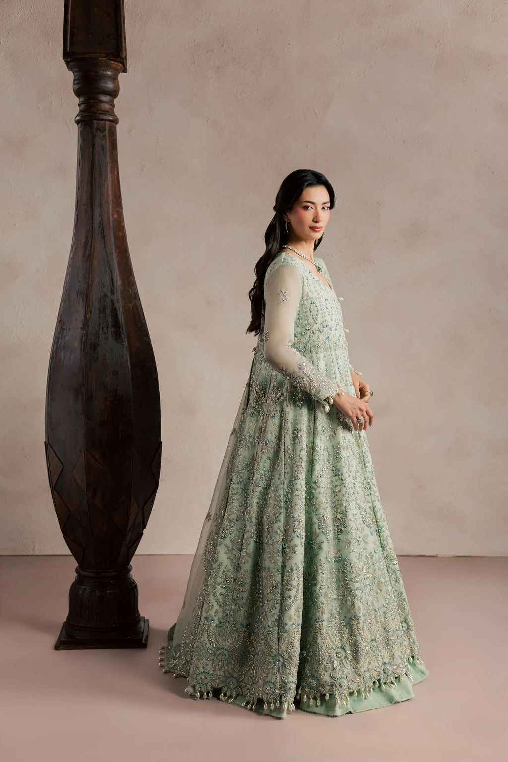 A woman wearing a long, light sage green embroidered gown stands beside a large dark wooden pillar against a soft beige studio backdrop.