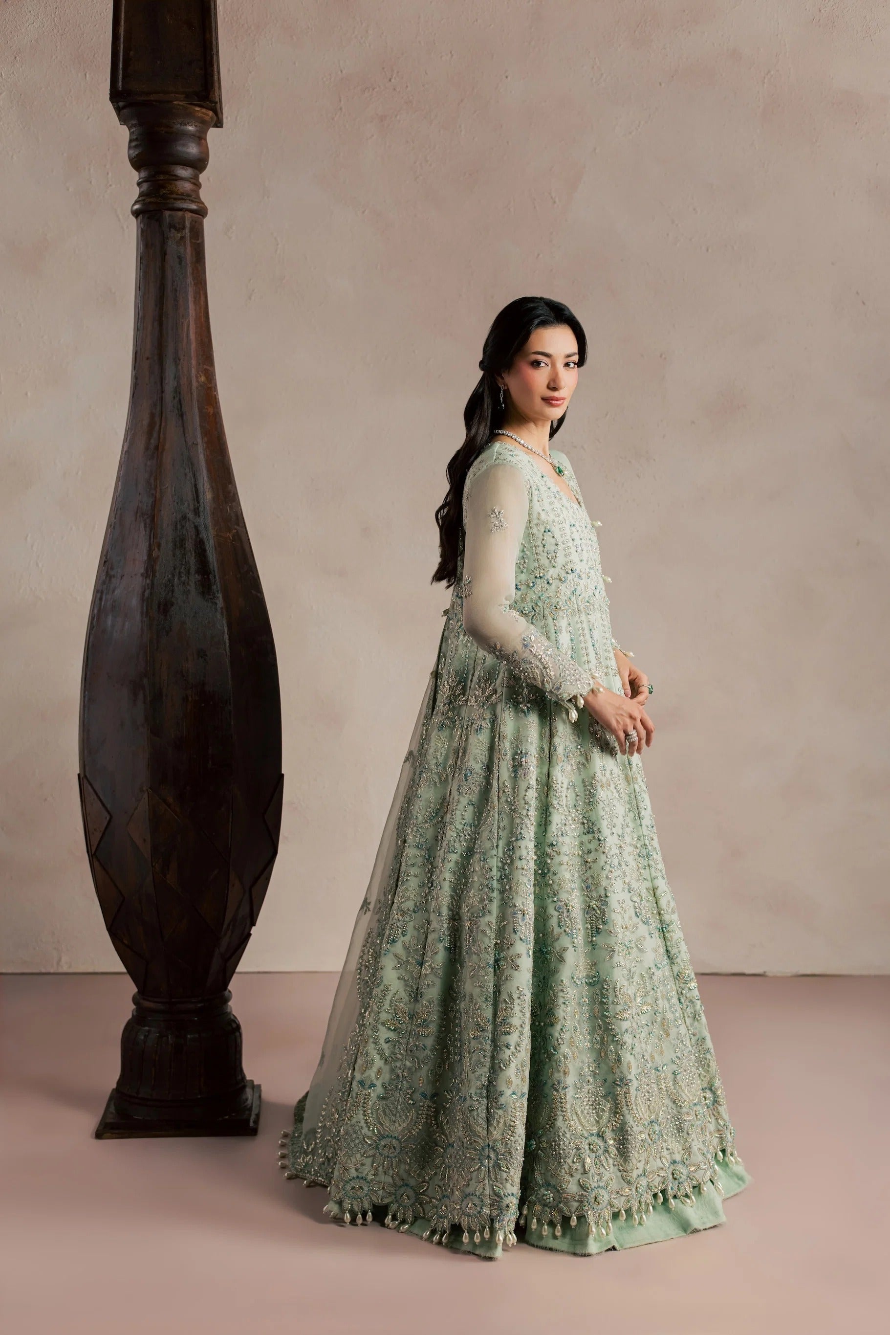 A woman wearing a long, light sage green embroidered gown stands beside a large dark wooden pillar against a soft beige studio backdrop.