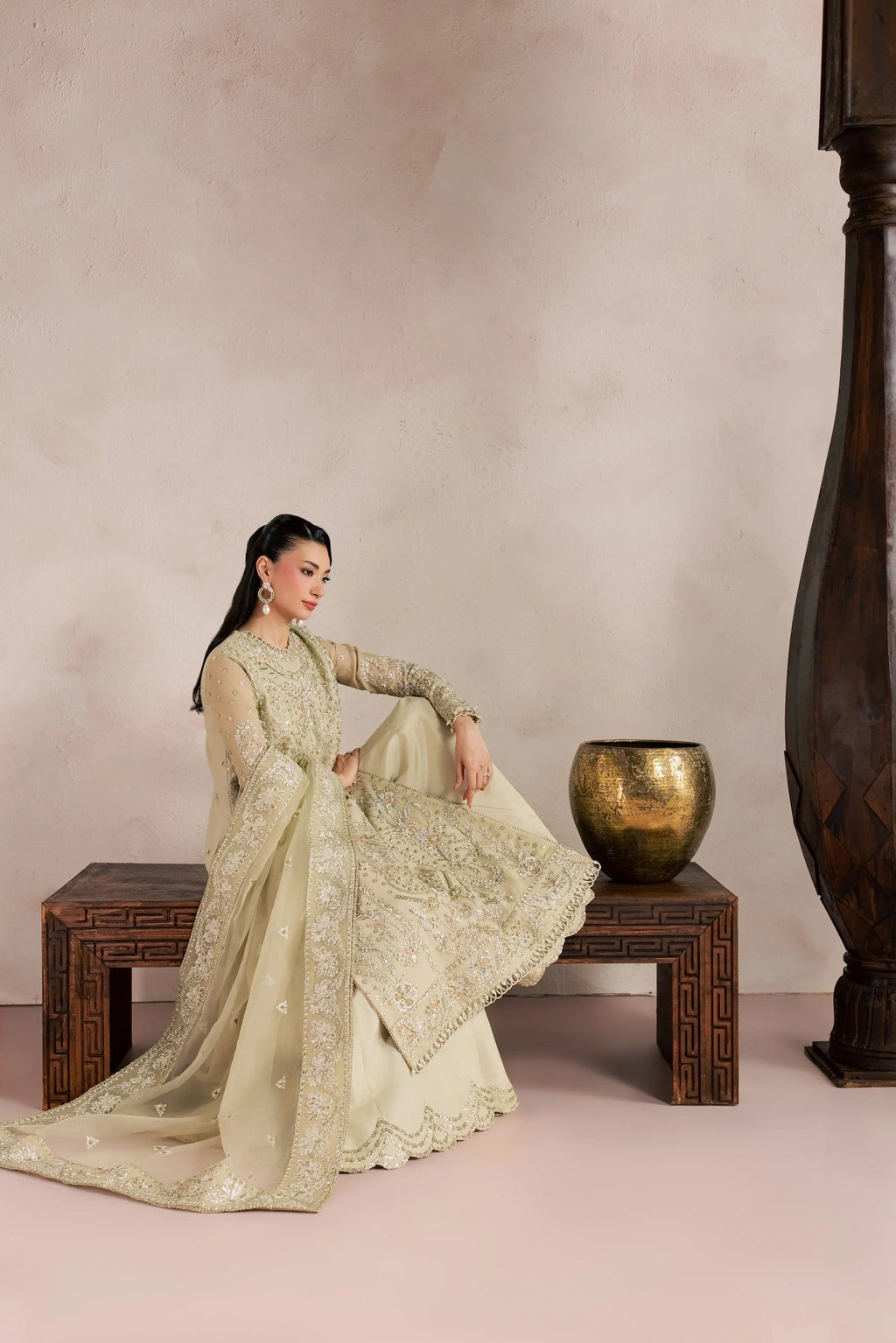 A woman models a traditional South Asian outfit, specifically a beige heavily embroidered kurta and matching dupatta, while sitting on a carved wooden bench in a styled studio setting.