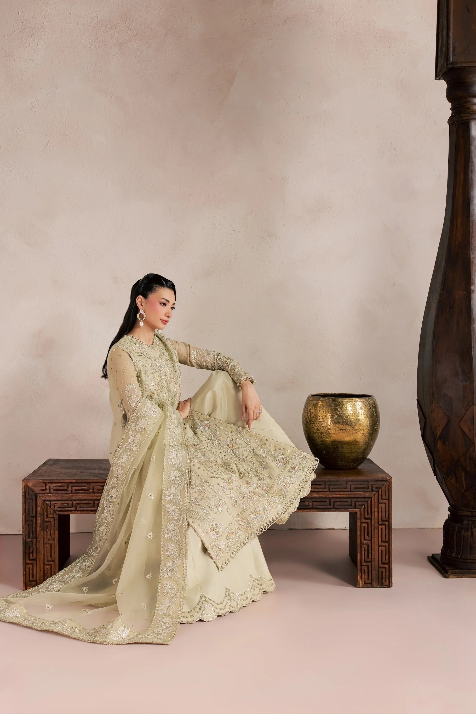 A woman models a traditional South Asian outfit, specifically a beige heavily embroidered kurta and matching dupatta, while sitting on a carved wooden bench in a styled studio setting.