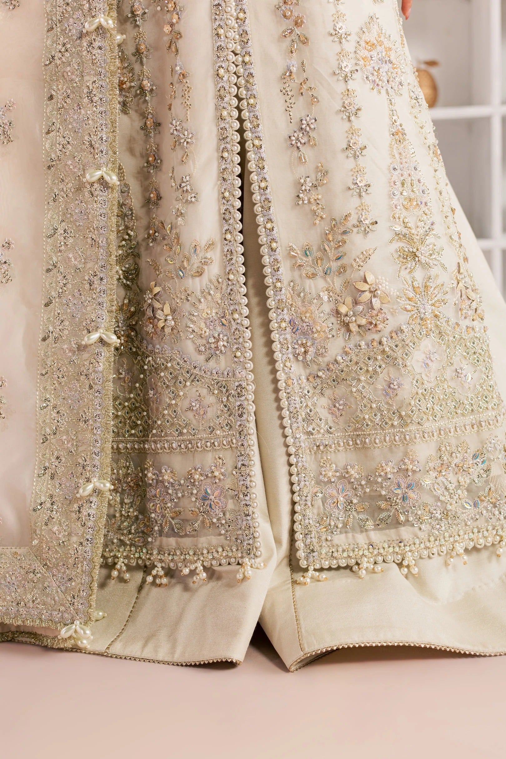 Close-up of an ornate cream-colored embroidered garment, likely a traditional ethnic dress or skirt, featuring intricate floral beadwork, sequins, and metallic detailing.