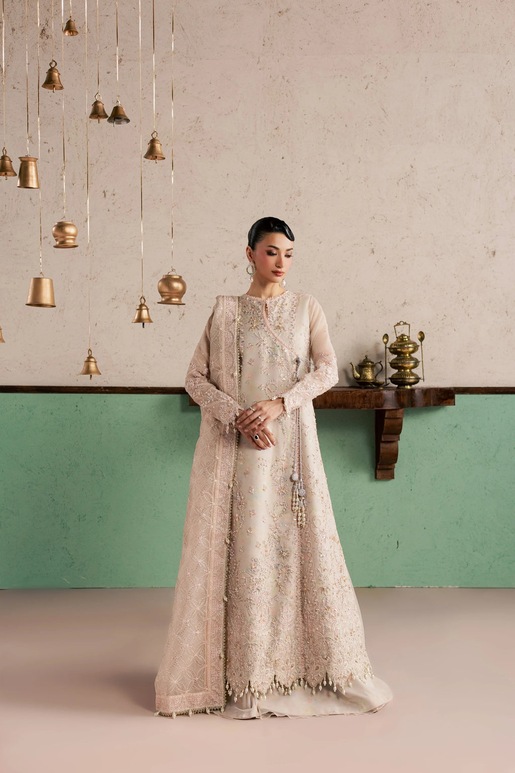 A woman stands indoors wearing a long, heavily embroidered light beige or champagne gown with sheer sleeves and a high neckline. The room has a textured light wall, a green lower wall section, hanging brass bell decorations, and a small wooden shelf with decorative objects.