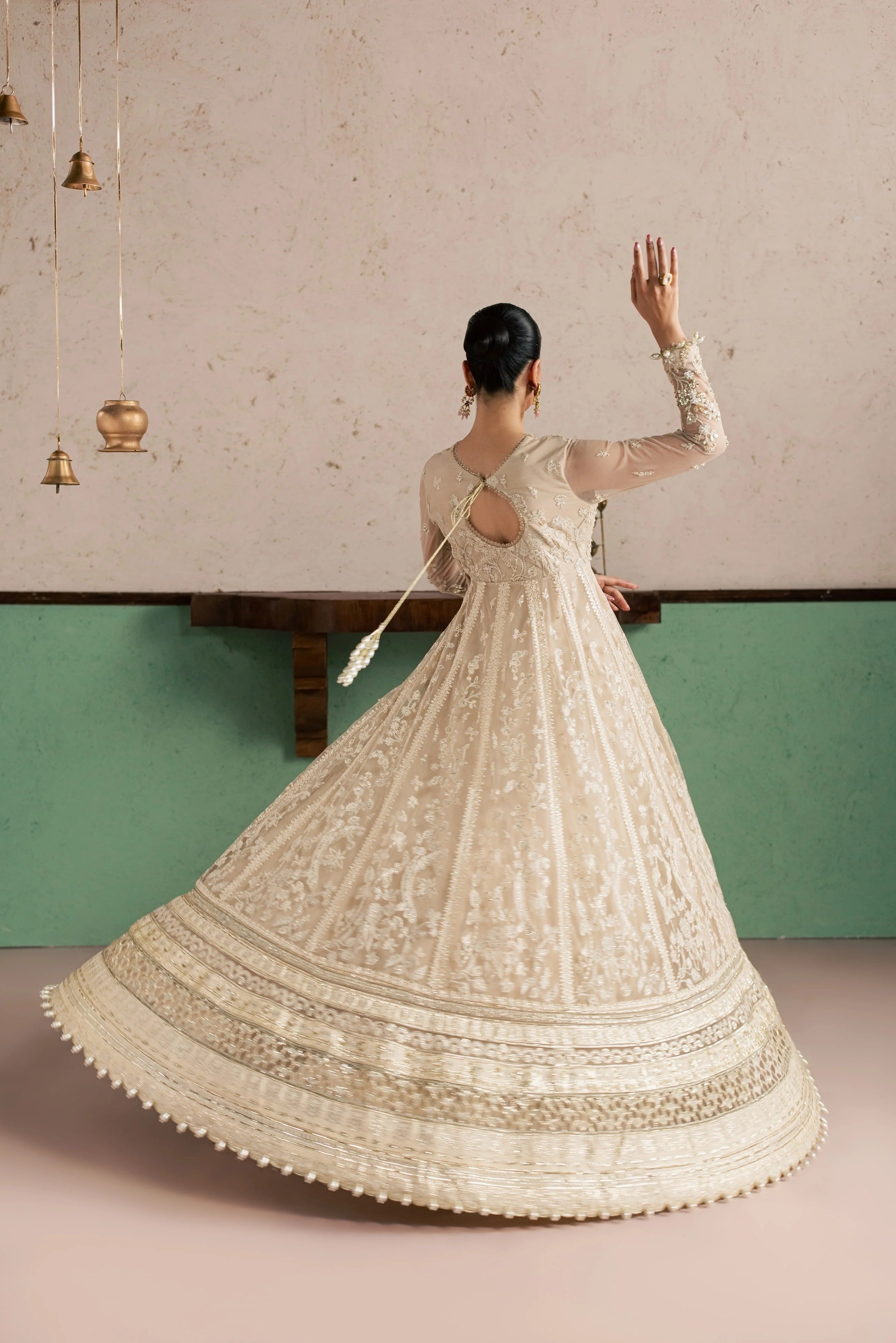A woman wearing a cream-colored heavily embroidered Anarkali gown, photographed from behind as she twirls to show the full flare of the skirt. The dress features intricate floral embroidery, a keyhole back with a tassel detail, and a heavily embellished border on the skirt.