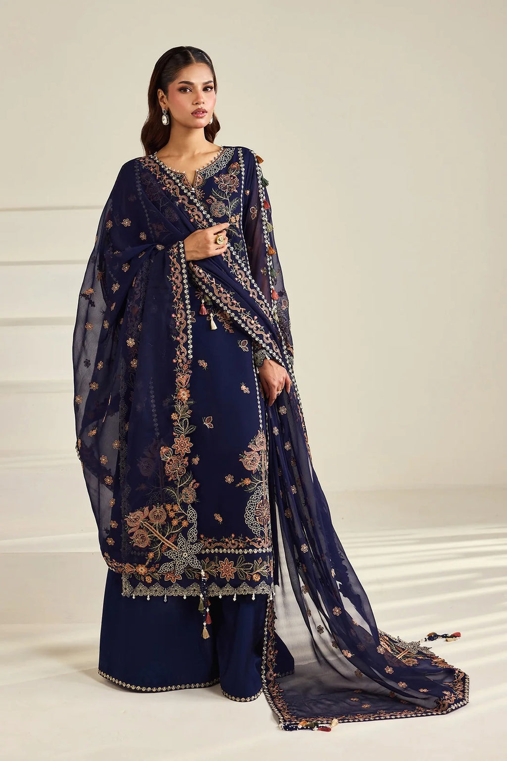 A woman models a navy blue traditional Indian outfit with intricate gold and beige floral embroidery, paired with a matching sheer dupatta, against a neutral cream-colored background.