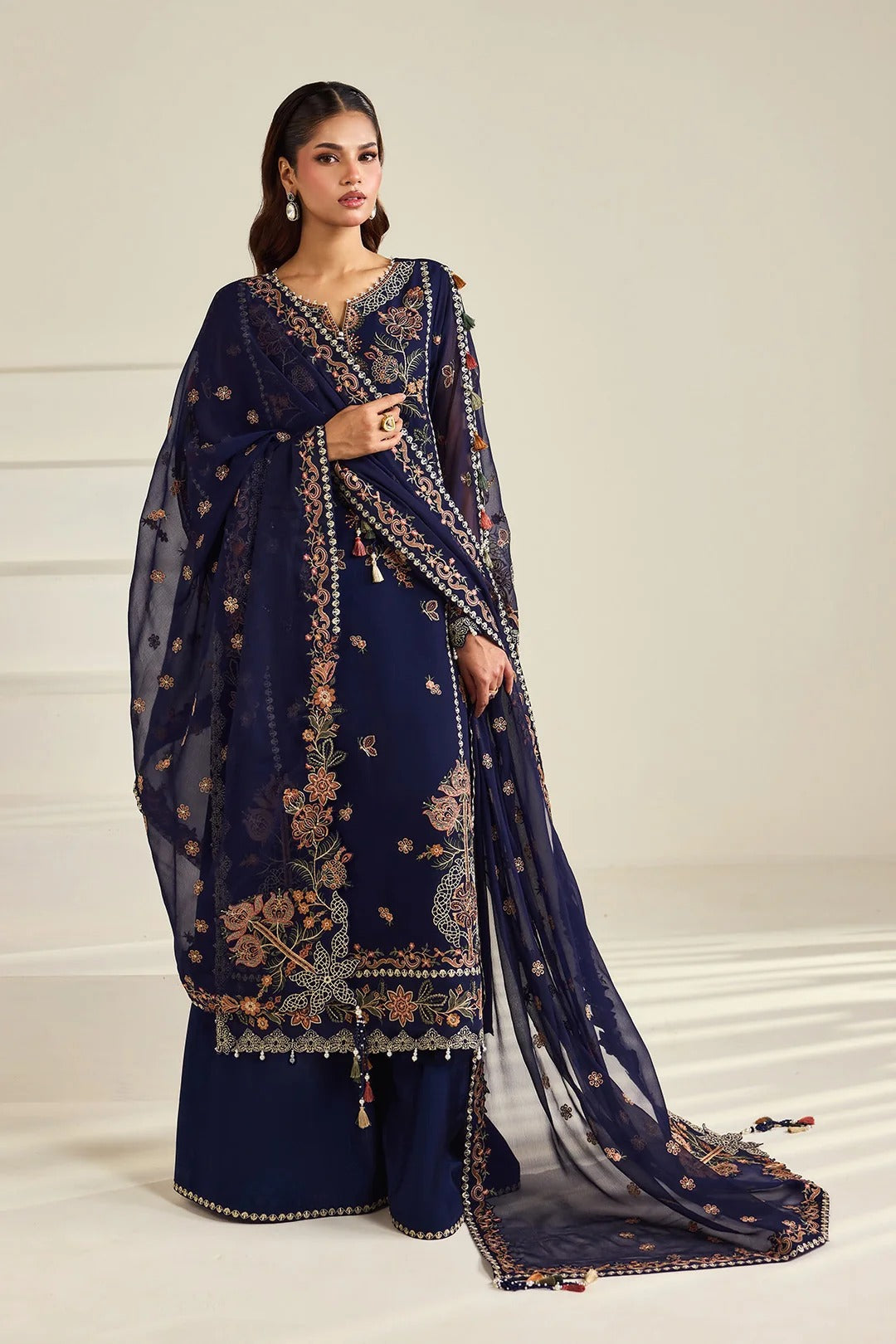 A woman models a navy blue traditional Indian outfit with intricate gold and beige floral embroidery, paired with a matching sheer dupatta, against a neutral cream-colored background.