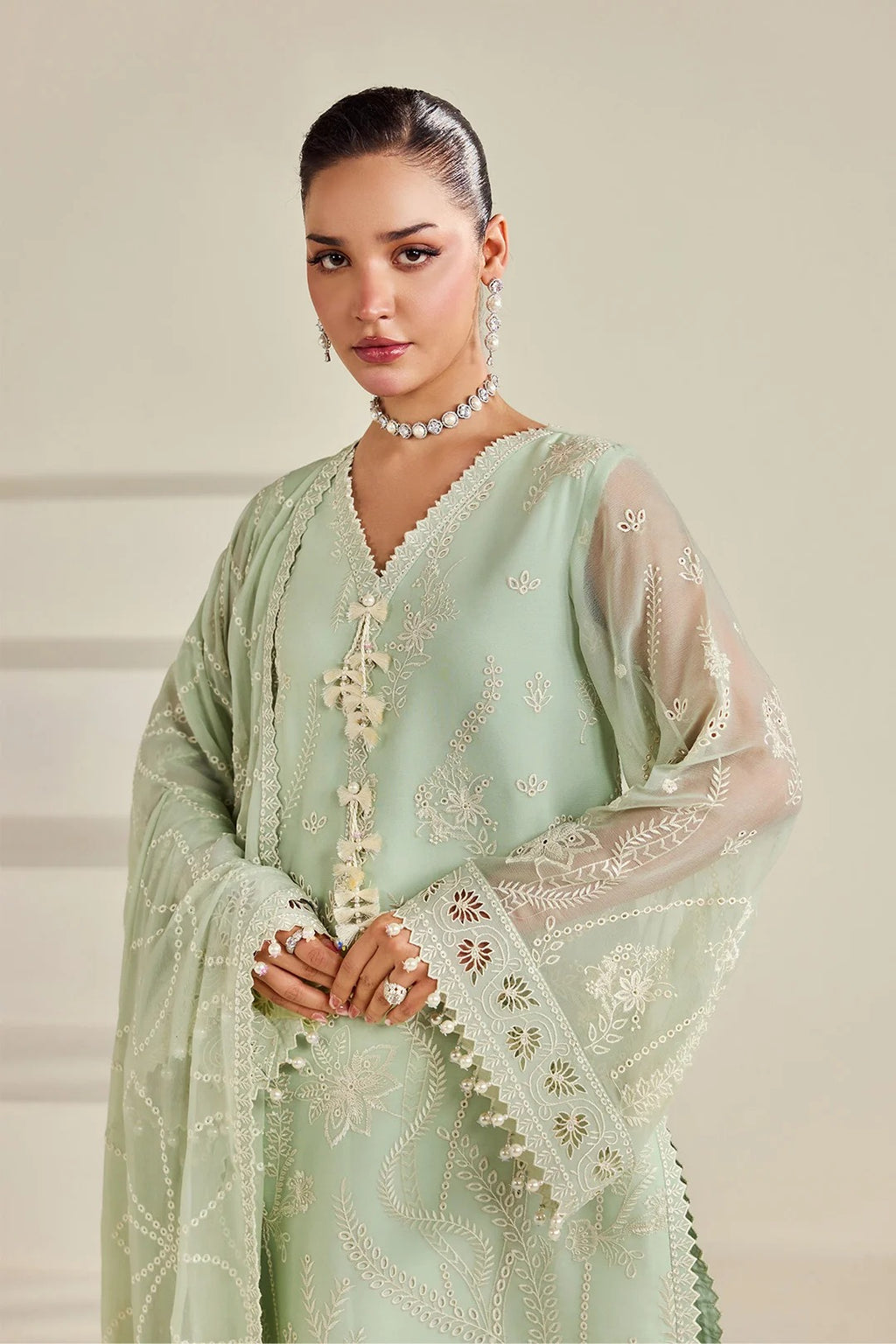 A woman modeling a light sage green traditional outfit with intricate white embroidery, sheer long sleeves, and a matching dupatta against a soft beige background.