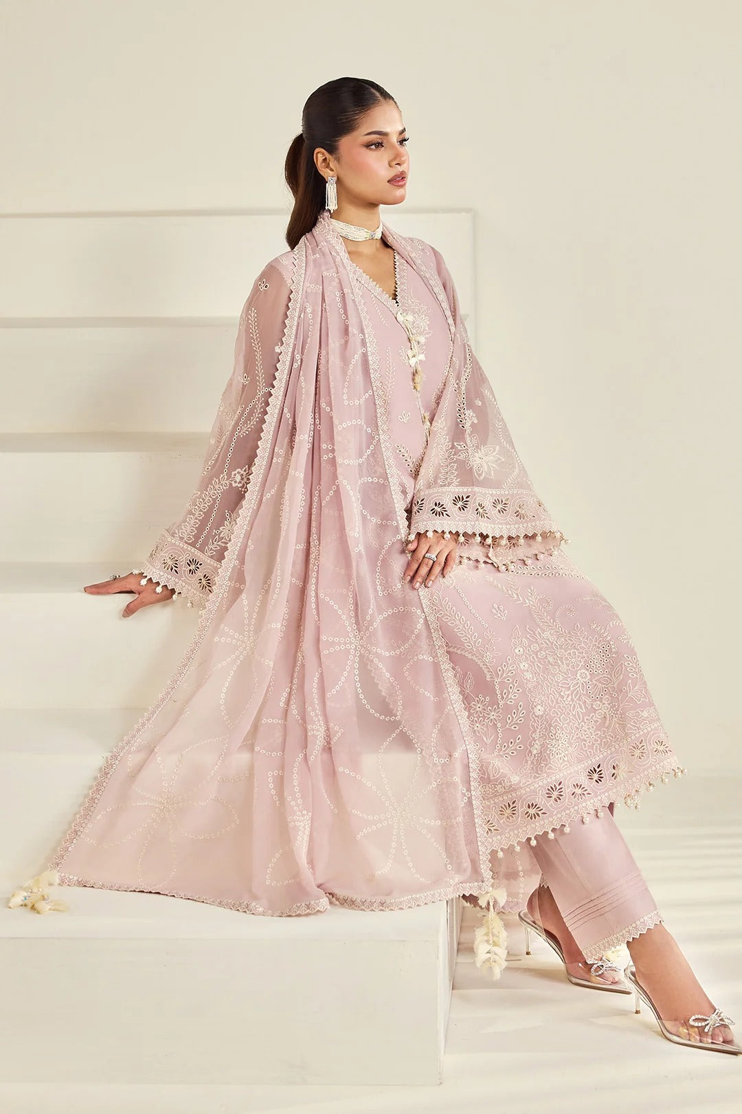 A woman models a light pink traditional outfit with a matching sheer dupatta draped over white steps. The outfit features intricate white embroidery and tassel details.
