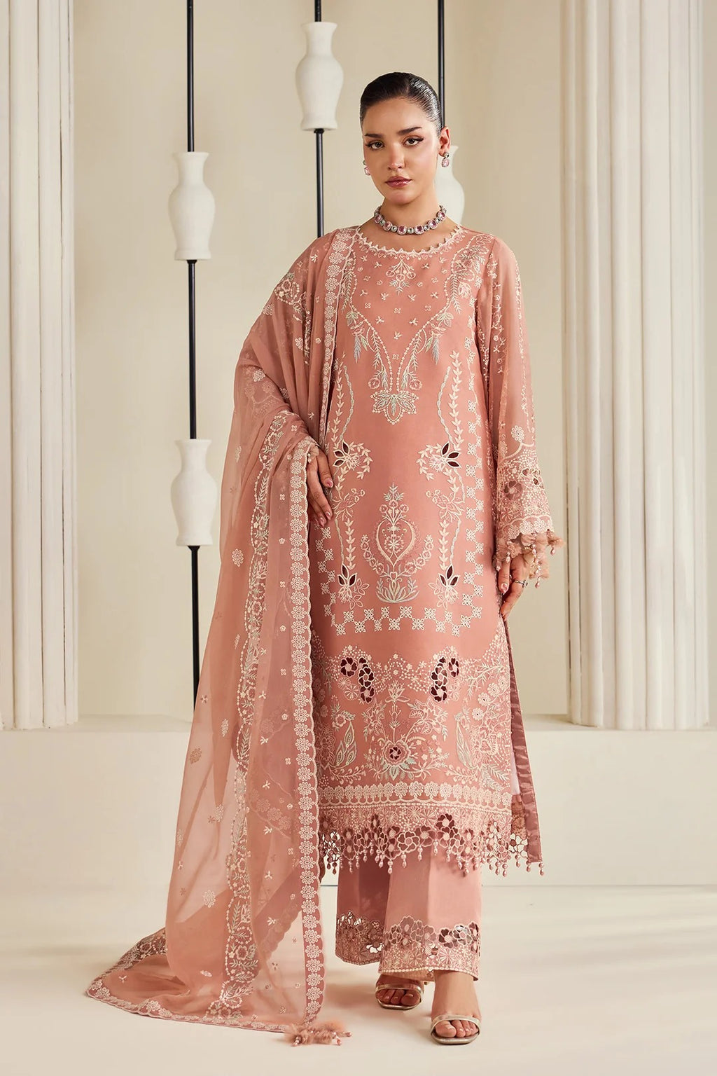 A woman models a traditional three-piece peach pink ethnic suit with intricate silver and white embroidery, featuring a long kurta, matching pants, and a sheer dupatta, set against a warm beige background with decorative columns.