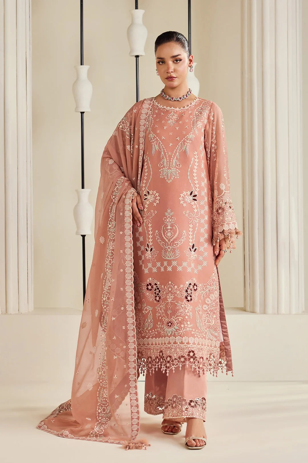 A woman models a traditional three-piece peach pink ethnic suit with intricate silver and white embroidery, featuring a long kurta, matching pants, and a sheer dupatta, set against a warm beige background with decorative columns.
