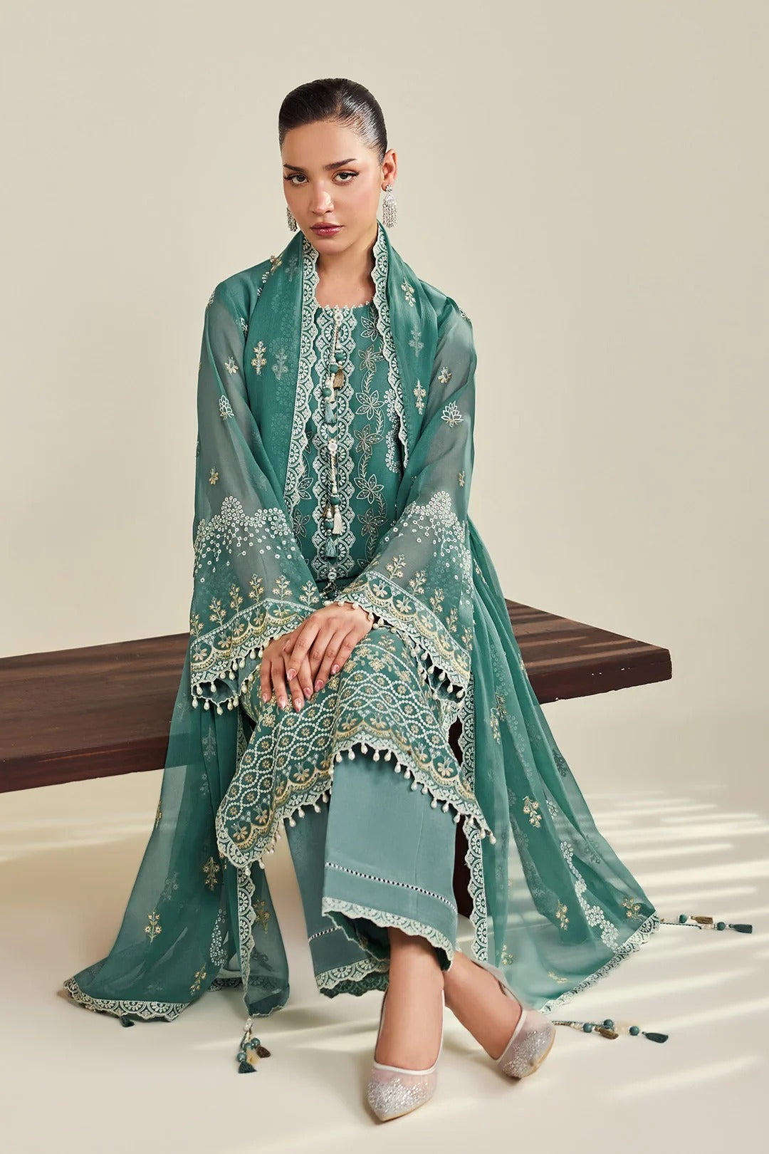A woman models a traditional three-piece green ethnic suit set consisting of a printed kurta, matching trousers, and a sheer embroidered dupatta, seated on a wooden bench against a neutral beige background.