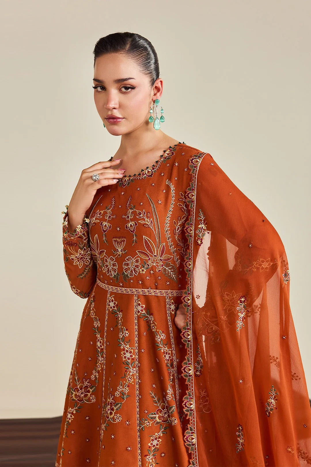 A woman models a rust-orange embroidered Anarkali gown with a matching sheer dupatta, set against a solid beige studio background.