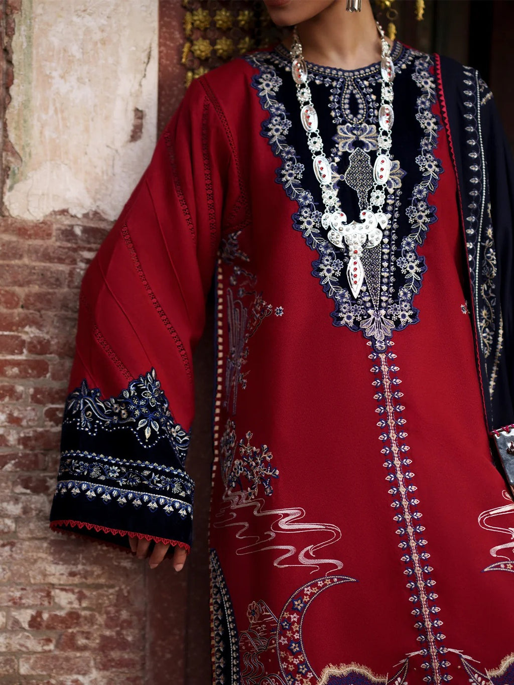 A close-up of a person wearing a rich red traditional embroidered tunic with navy and silver patterns, standing against a rustic brick wall.
