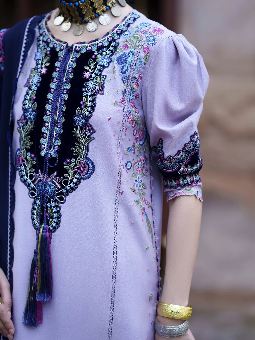 A close-up of a lavender traditional embroidered tunic with intricate multicolor floral embroidery on the neckline and sleeves, worn by a model with gold jewelry.