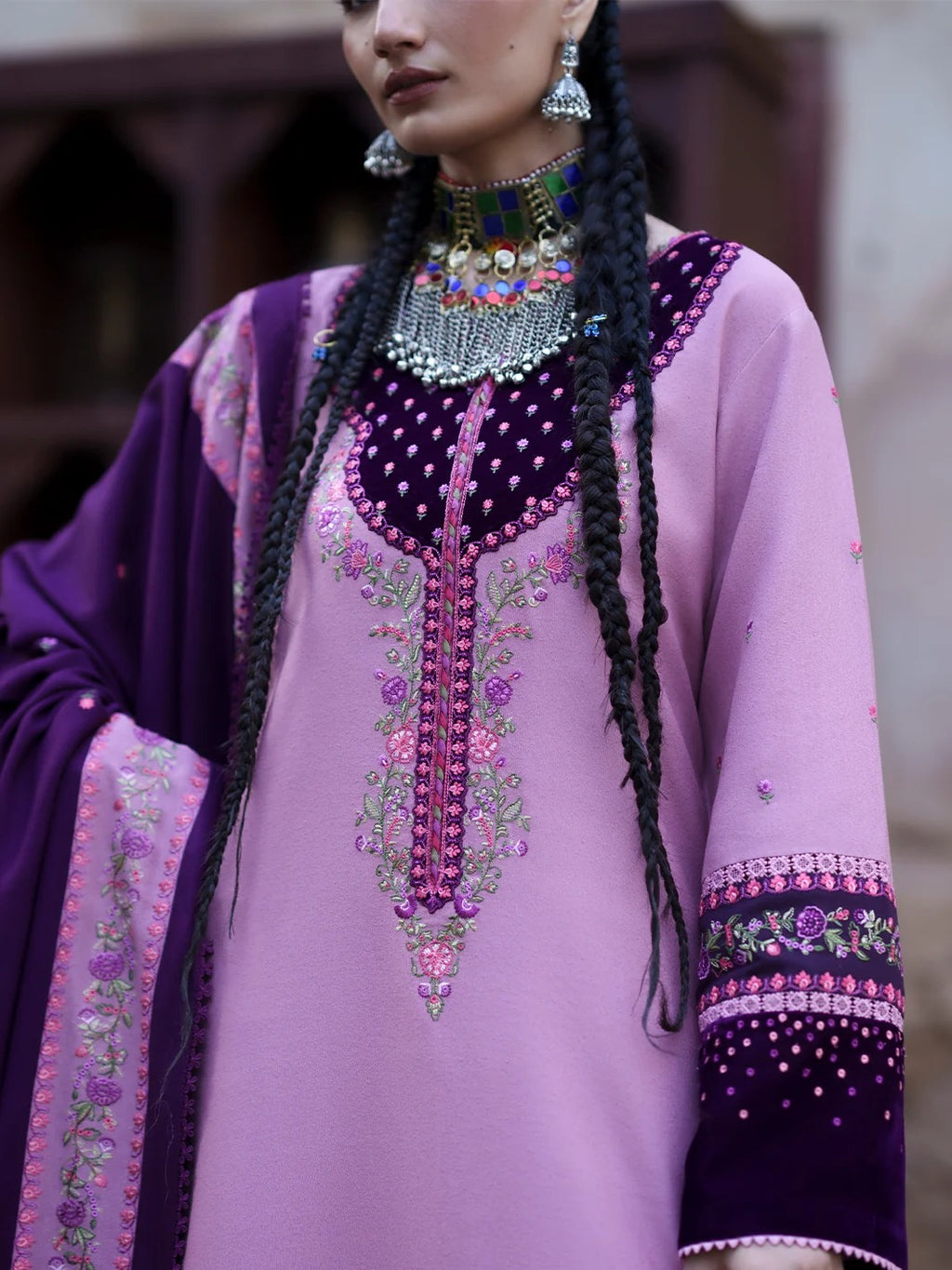 A close-up of a model wearing a traditional purple and lavender embroidered outfit with intricate floral embroidery, a matching purple dupatta, and ornate silver jewelry.