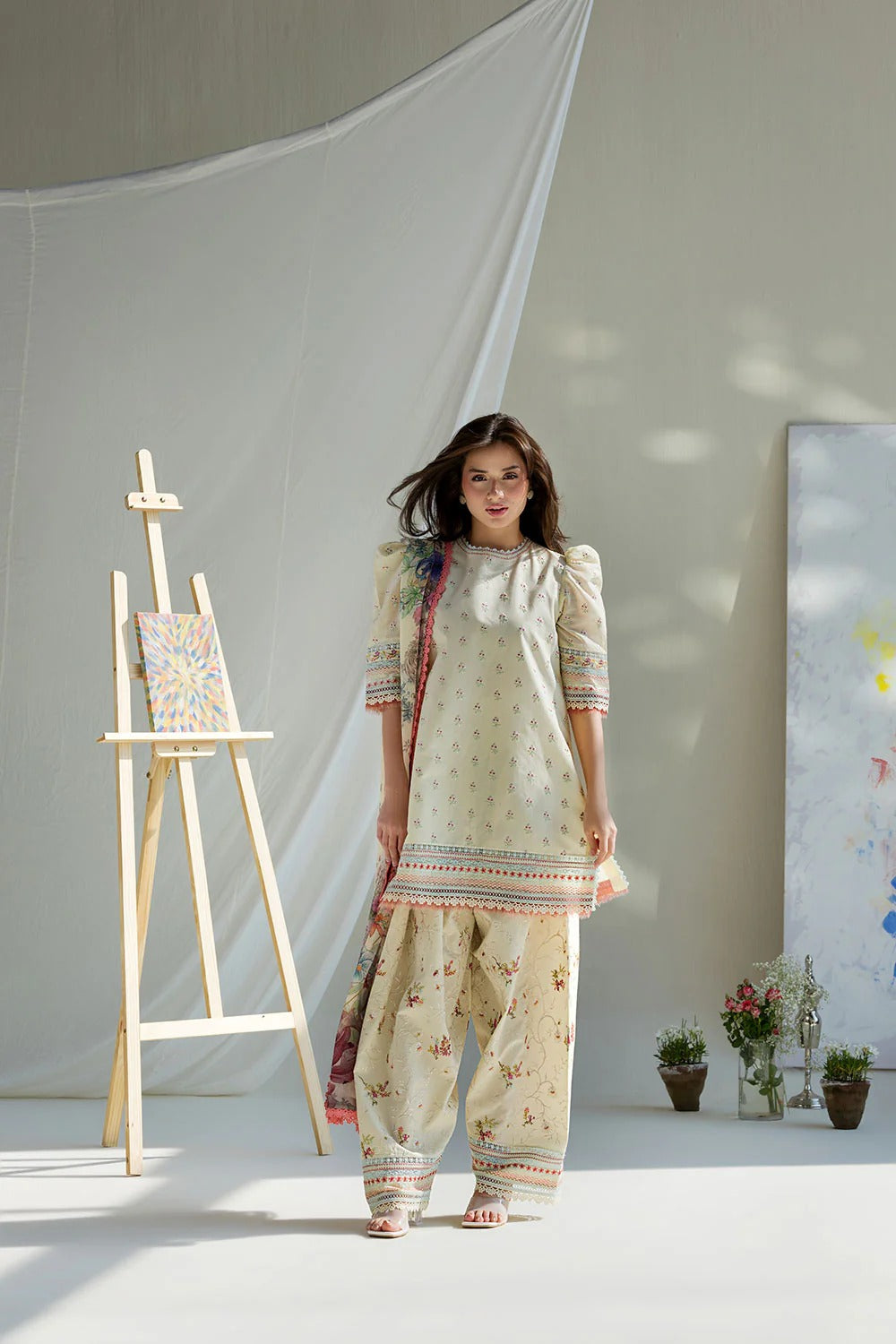 Woman in a traditional outfit standing in a room with an easel and plants.