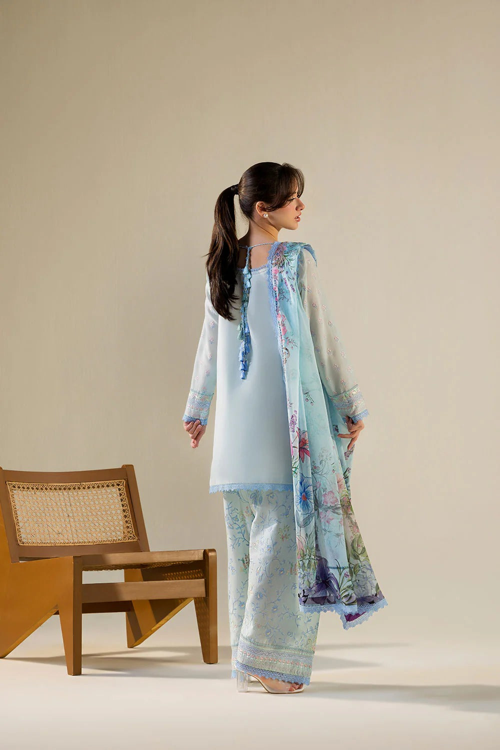 Woman in a light blue traditional outfit with a floral dupatta standing next to a wooden chair against a beige background