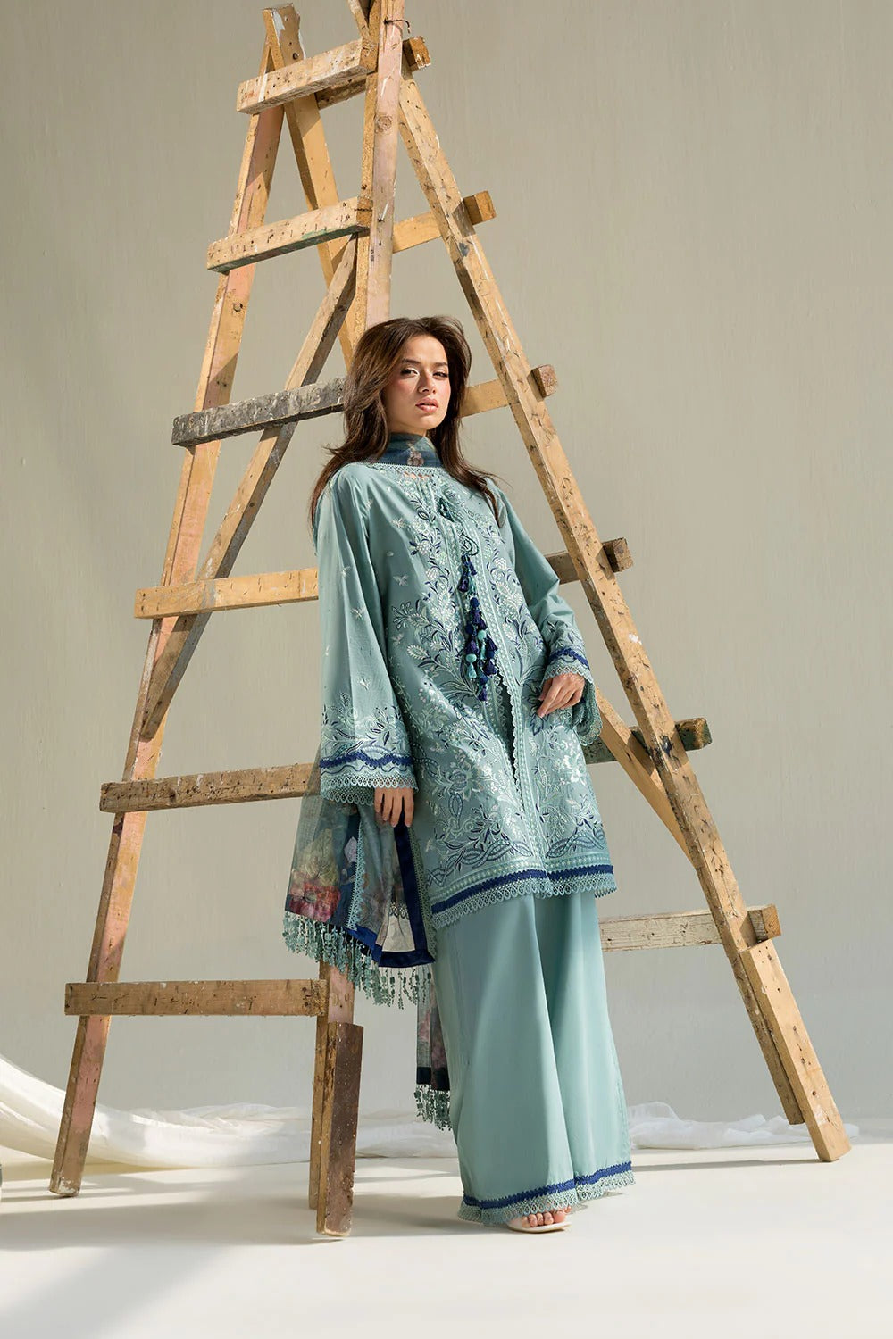 Woman in a light blue traditional outfit standing next to a wooden ladder on a plain background