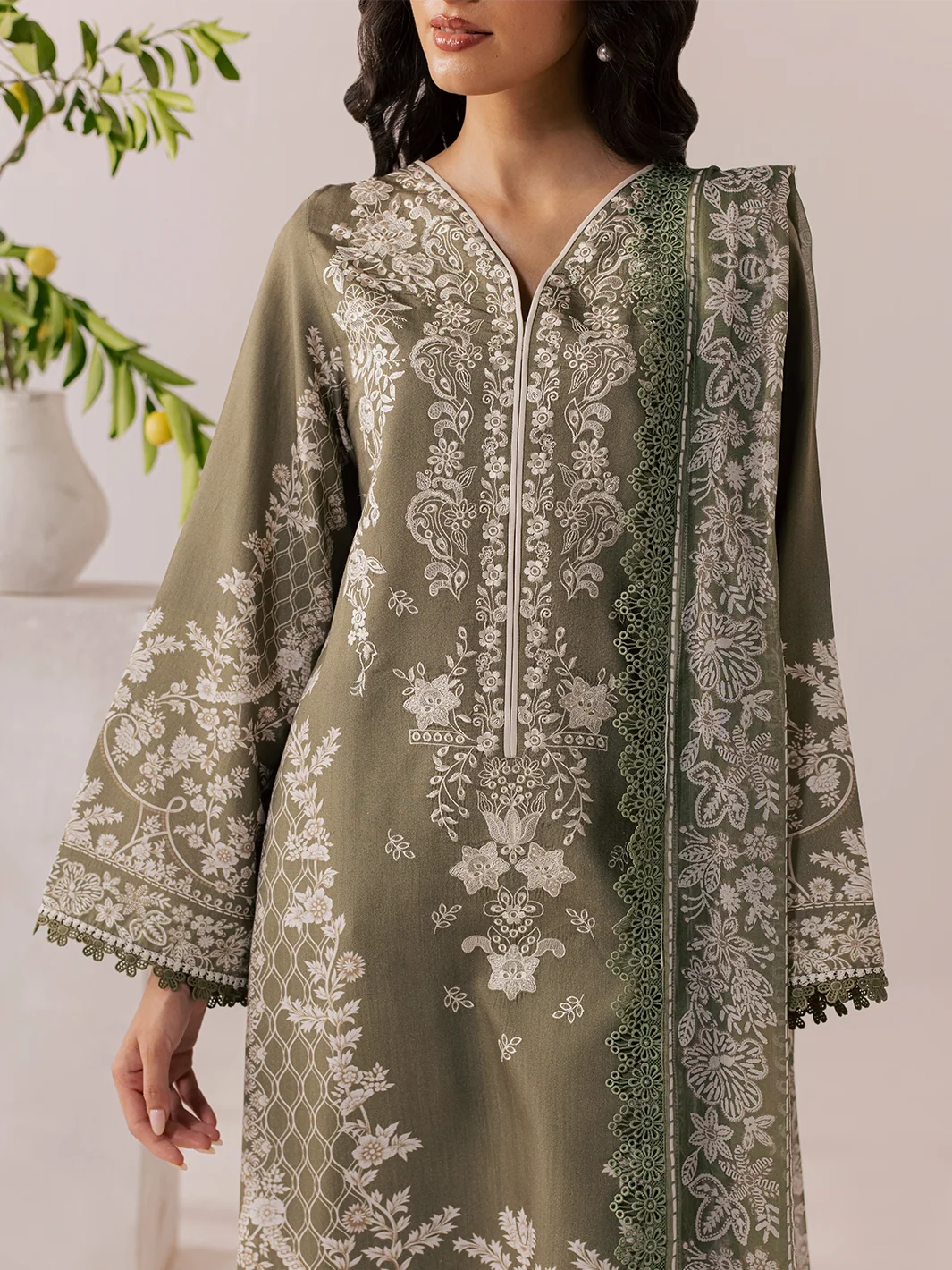 A woman models a sage green embroidered kurta with intricate white floral patterns and a matching embroidered dupatta, set against a soft beige background with a decorative plant in the corner.