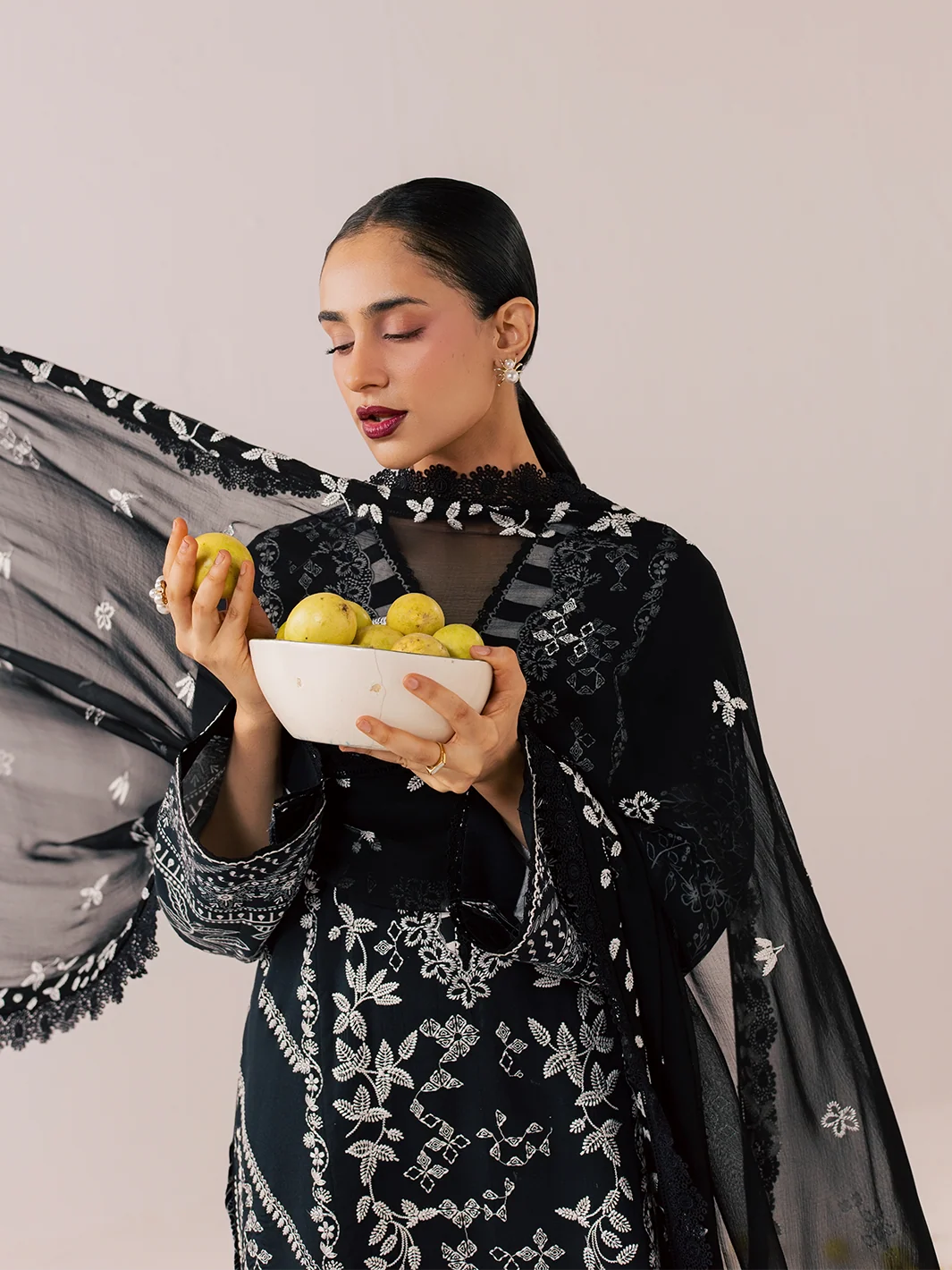 A woman models a black embroidered traditional outfit with a matching sheer embroidered dupatta, holding a white bowl of lemons against a solid light beige background.