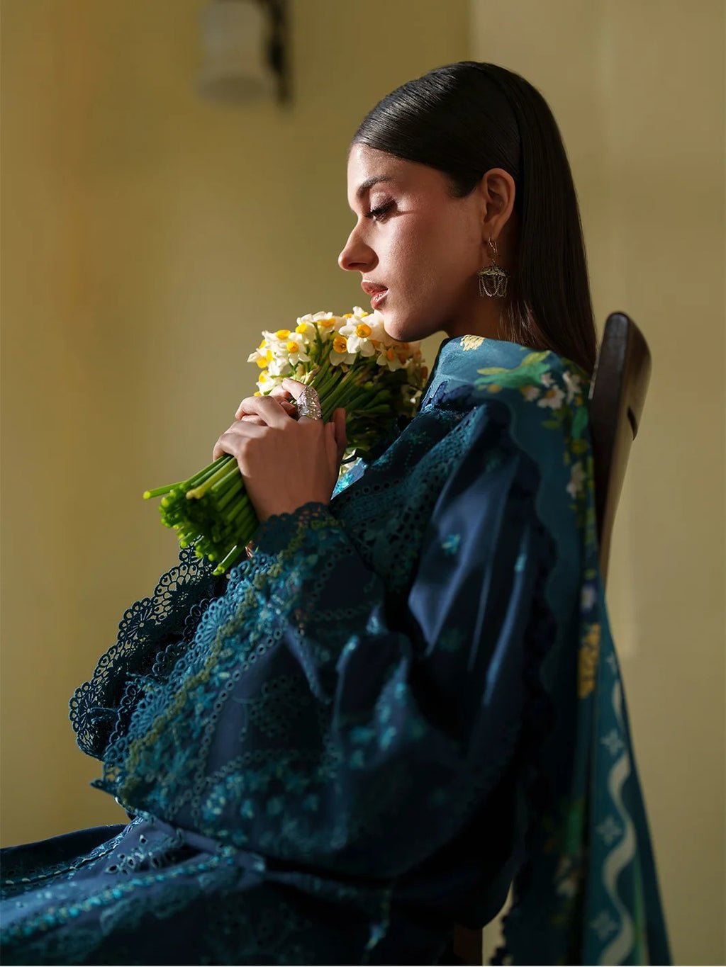 Woman holding a bouquet of flowers, wearing a blue floral dress, sitting against a plain background.