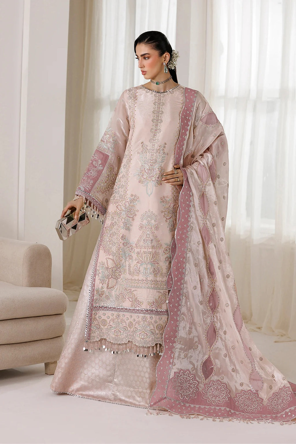 A woman models a traditional three-piece salwar kameez suit in a soft blush pink color with intricate silver and gold embroidery. The set includes a long kurta, matching trousers, and a sheer dupatta draped over her shoulder.