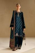 A woman models a dark teal embroidered traditional outfit with matching pants and a dupatta, holding a decorative black and gold clutch bag against a textured beige studio backdrop.