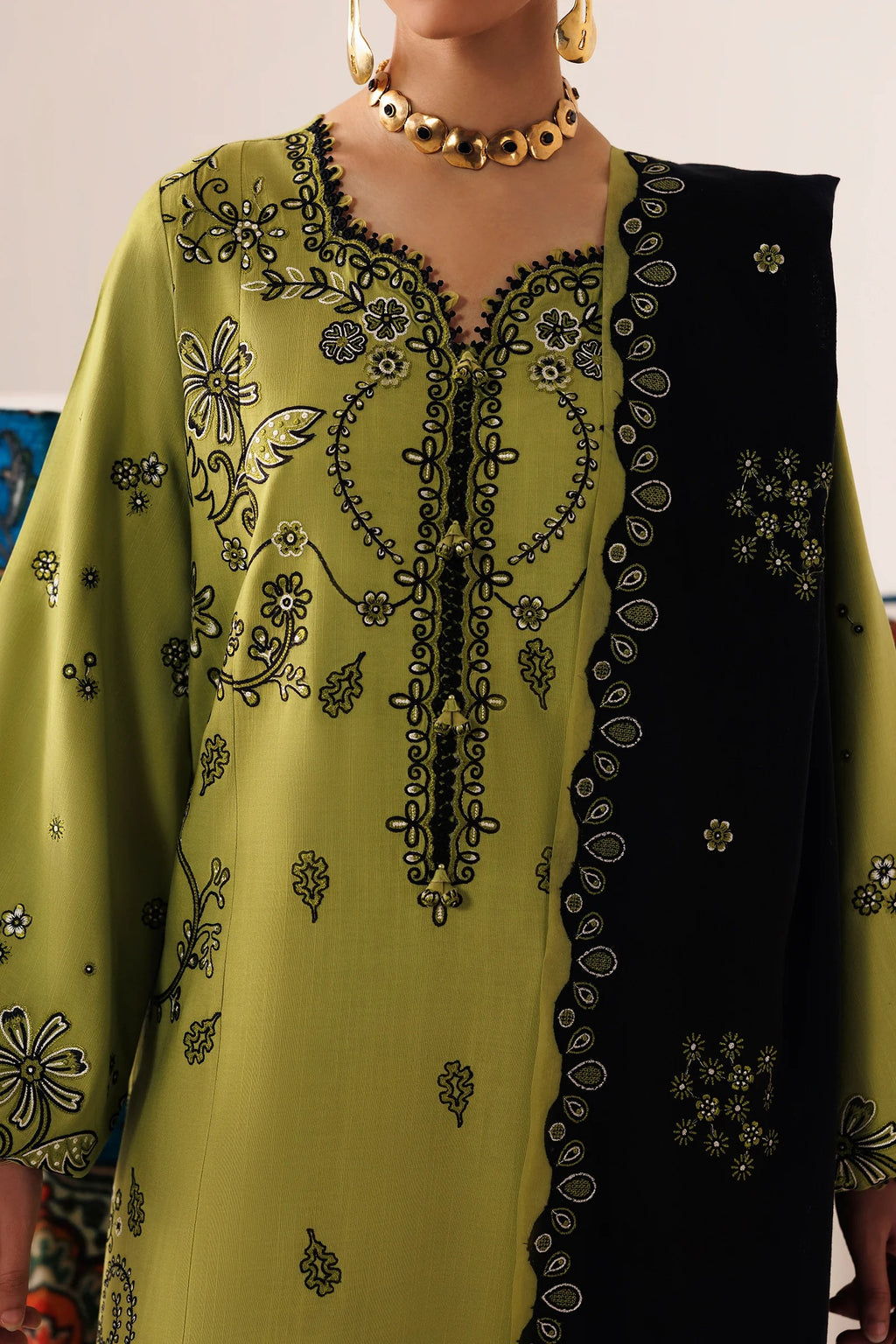 A close-up of a woman wearing a traditional embroidered kurta in olive green with black floral embroidery, paired with a black dupatta featuring gold and black border detailing, accessorized with gold jewelry.