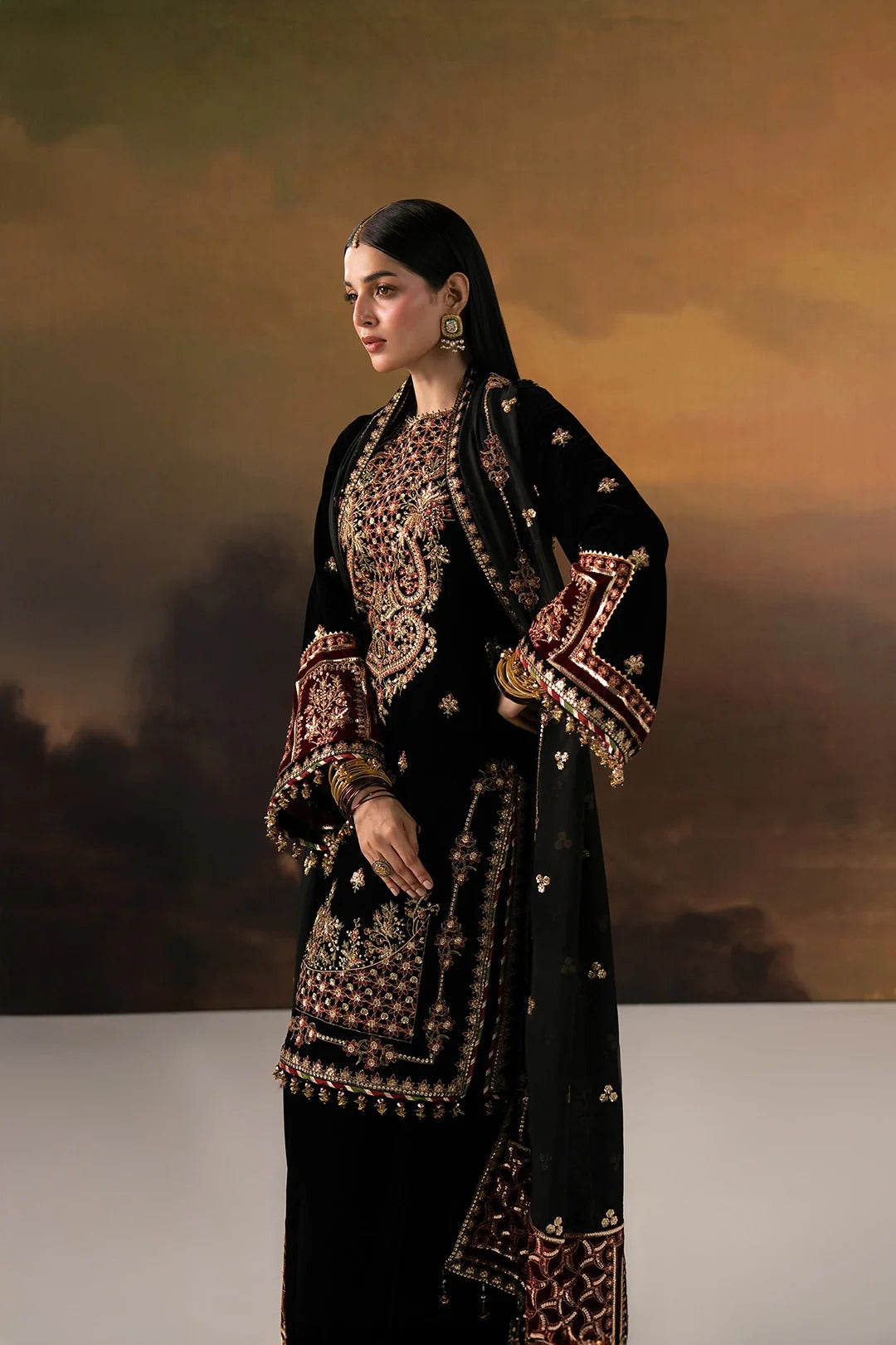 A woman models a traditional black embroidered kurta set with a matching dupatta against a warm, painterly brown background. The outfit features intricate gold and red floral embroidery.