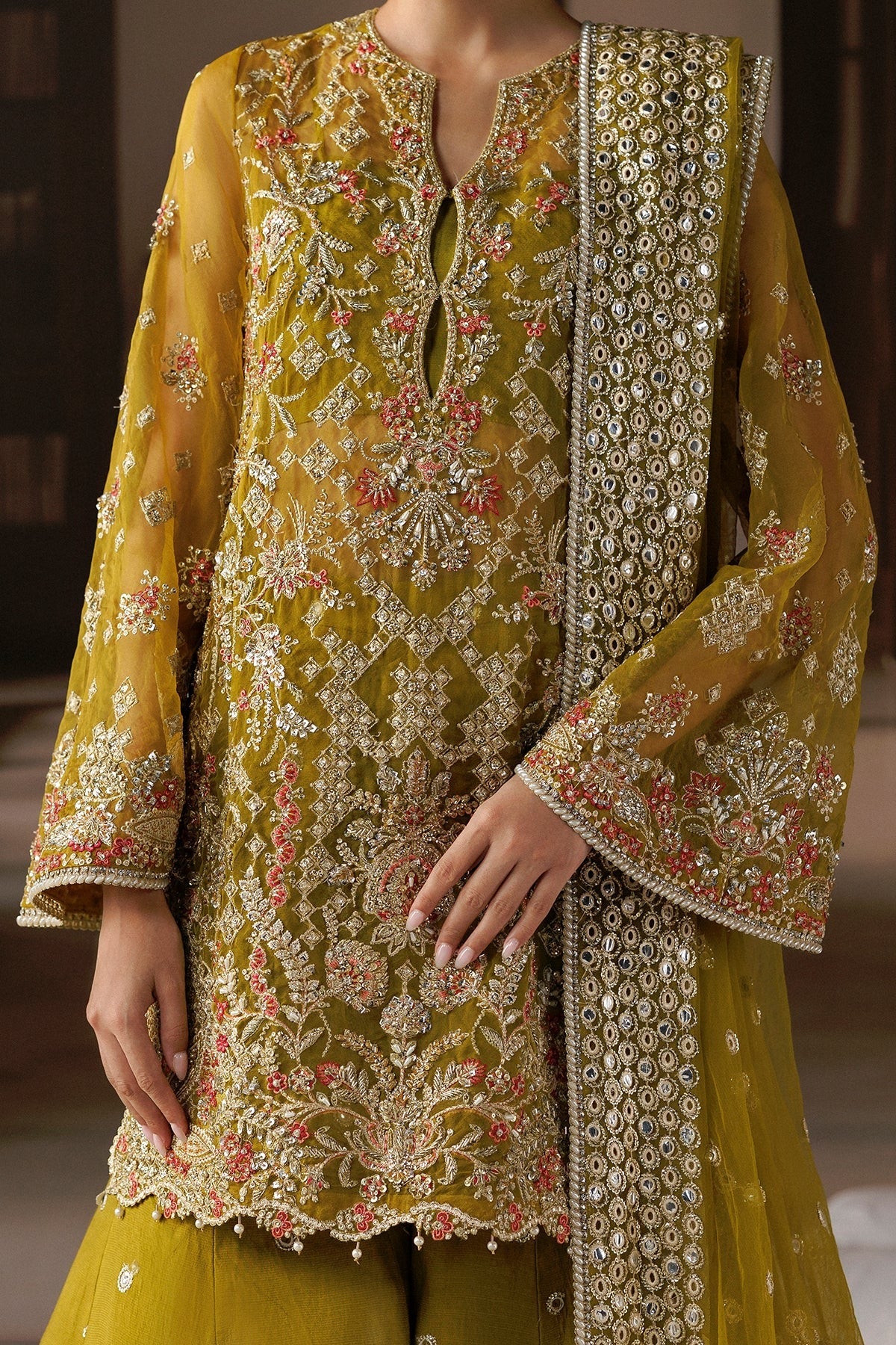 A close-up of a woman wearing an ornate olive-yellow embroidered traditional outfit with a matching dupatta. The kurta features intricate floral embroidery in gold, silver, and pink, with a split neckline and a decorative scalloped hem.