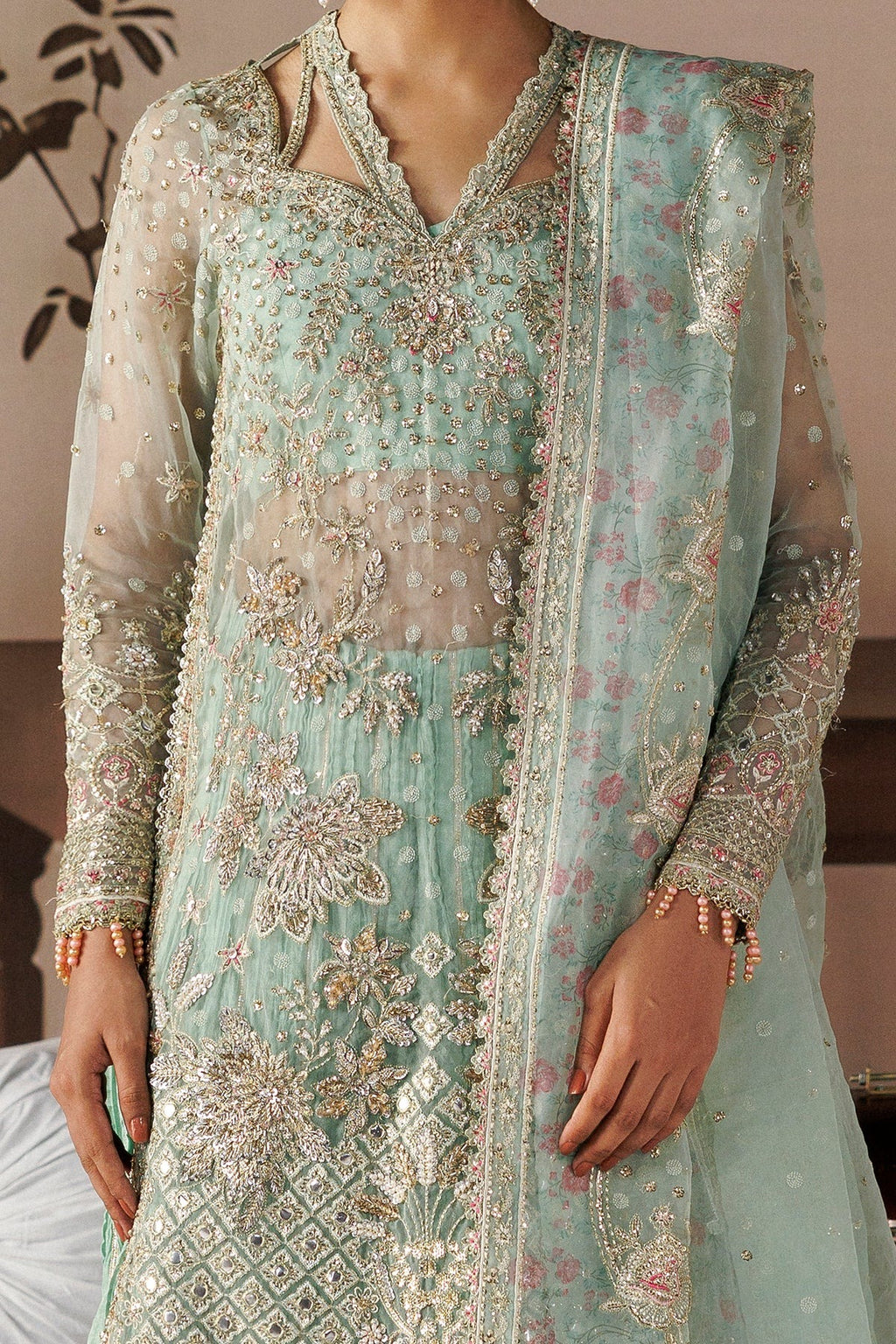 A close-up of a model wearing a mint green embroidered traditional outfit with a matching sheer dupatta. The outfit features intricate floral and geometric embroidery in metallic gold, silver, and pink on a light green base.