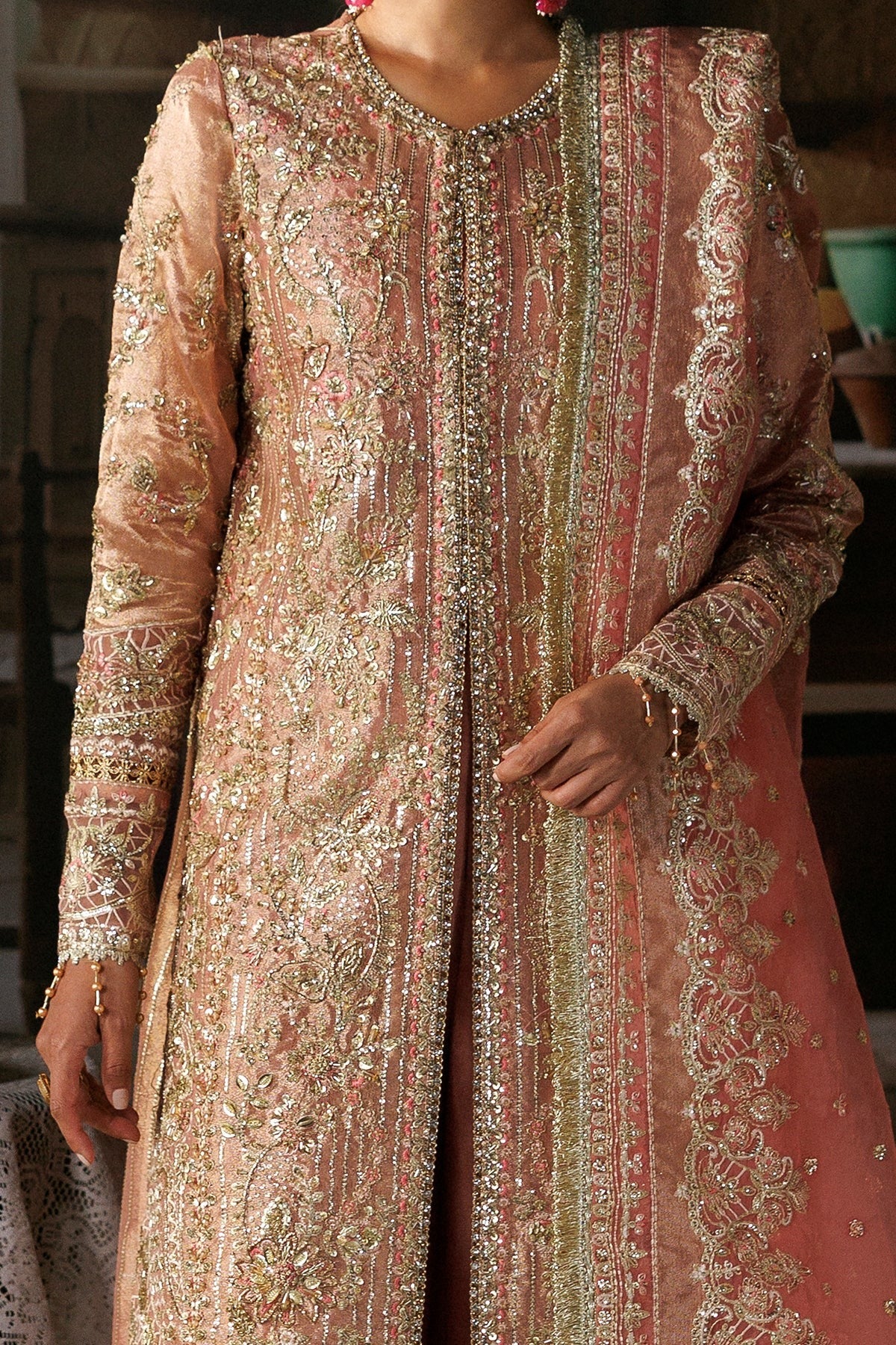 A close-up of a person wearing an ornate peach-pink traditional embroidered outfit with heavy gold metallic detailing and a matching sheer dupatta.
