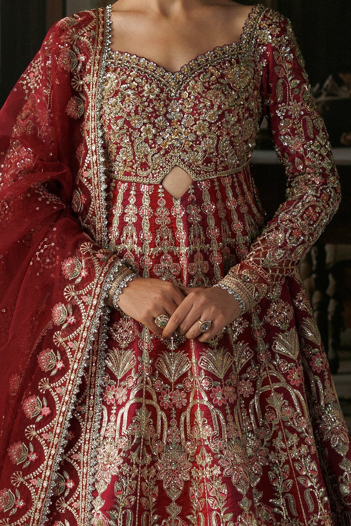 A close-up of a woman wearing an ornate red and gold embroidered traditional dress with a matching sheer dupatta, featuring intricate metallic embellishments and detailed patterns.