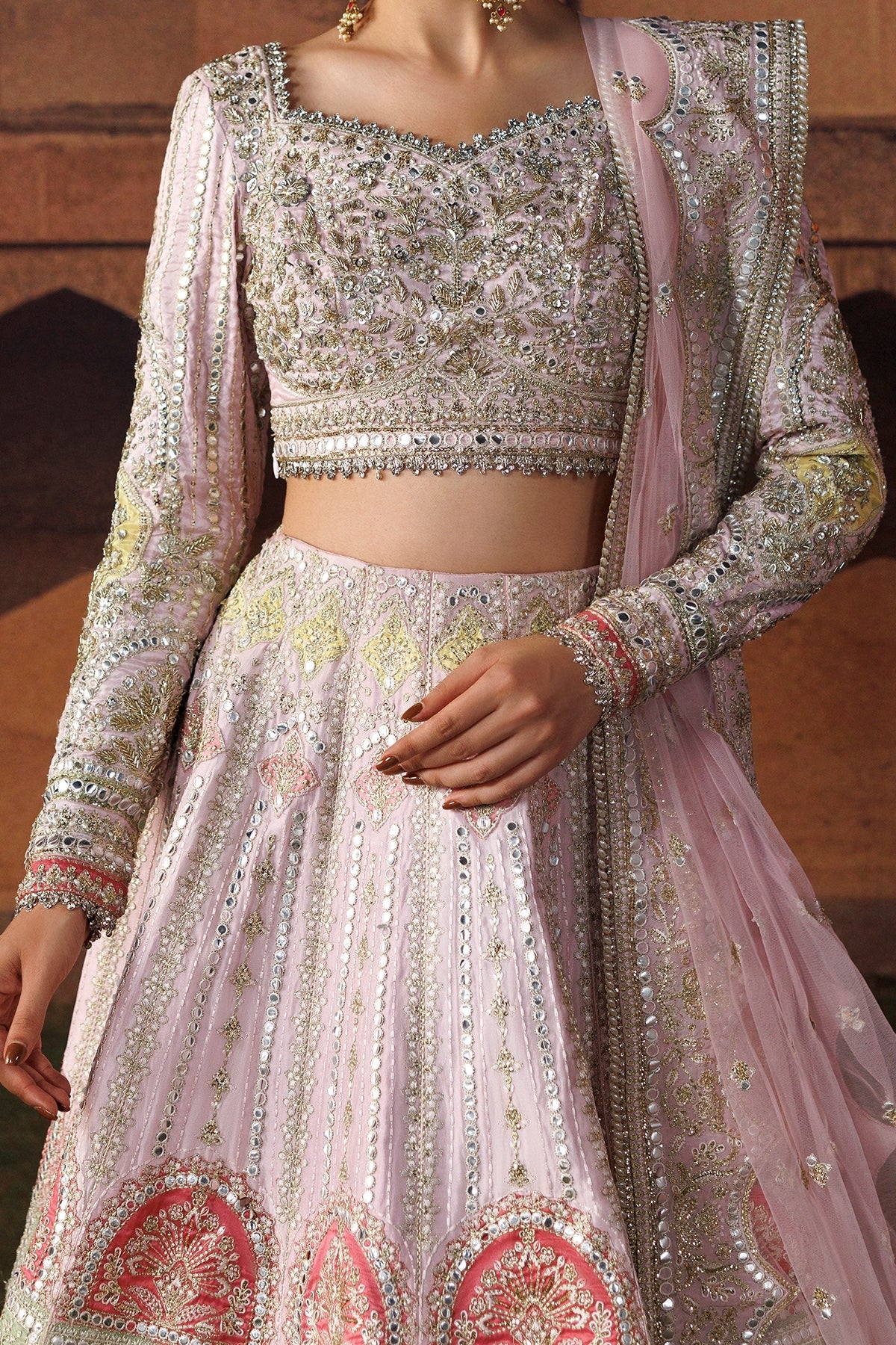 A close-up of a model wearing an ornate light pink traditional outfit with heavy silver and gold embroidery, a matching embroidered blouse, and a sheer pink dupatta draped over one shoulder.