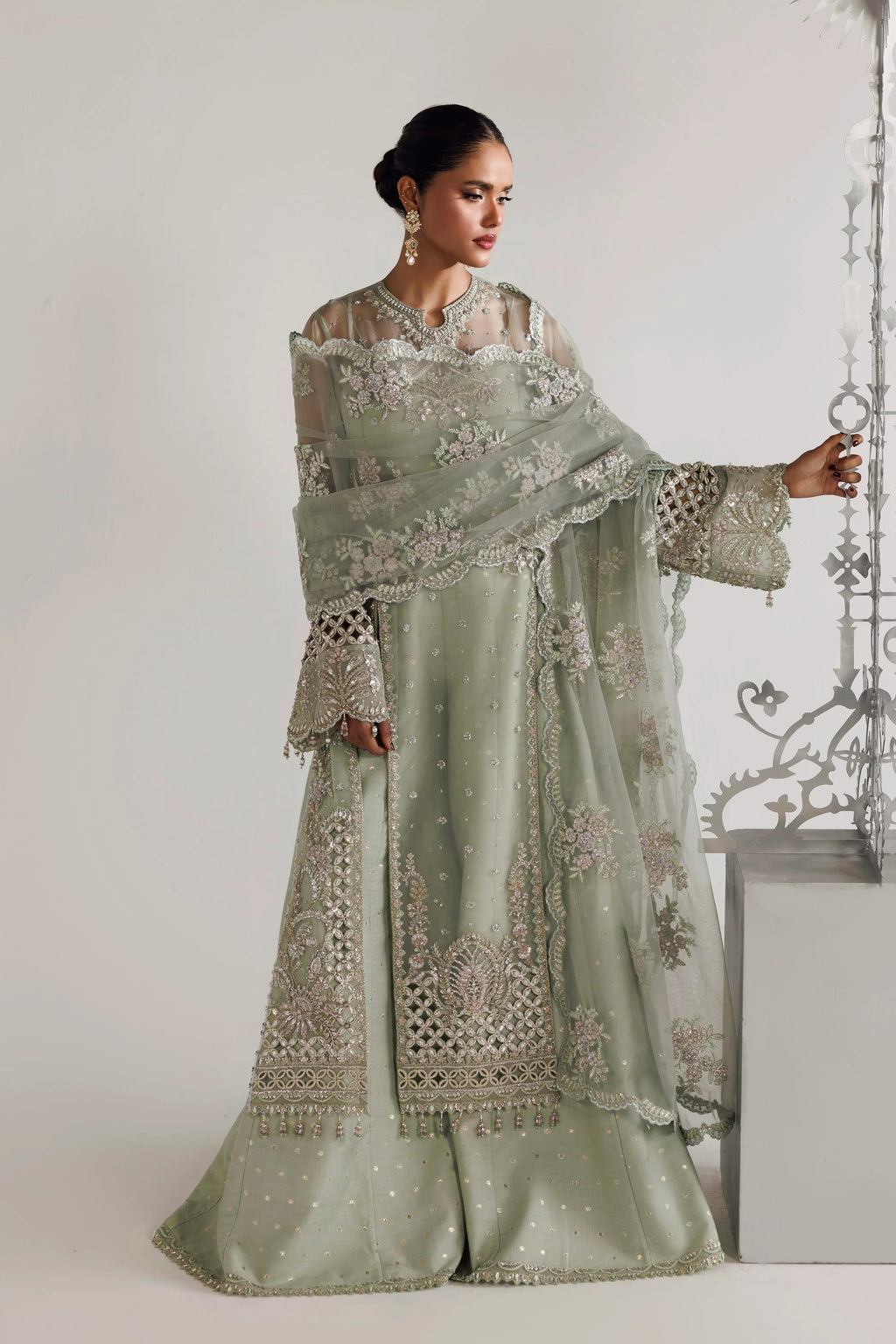 A woman models a light sage green embroidered traditional outfit with a matching dupatta, featuring intricate silver mirror work and floral embroidery, set against a minimalist studio background.