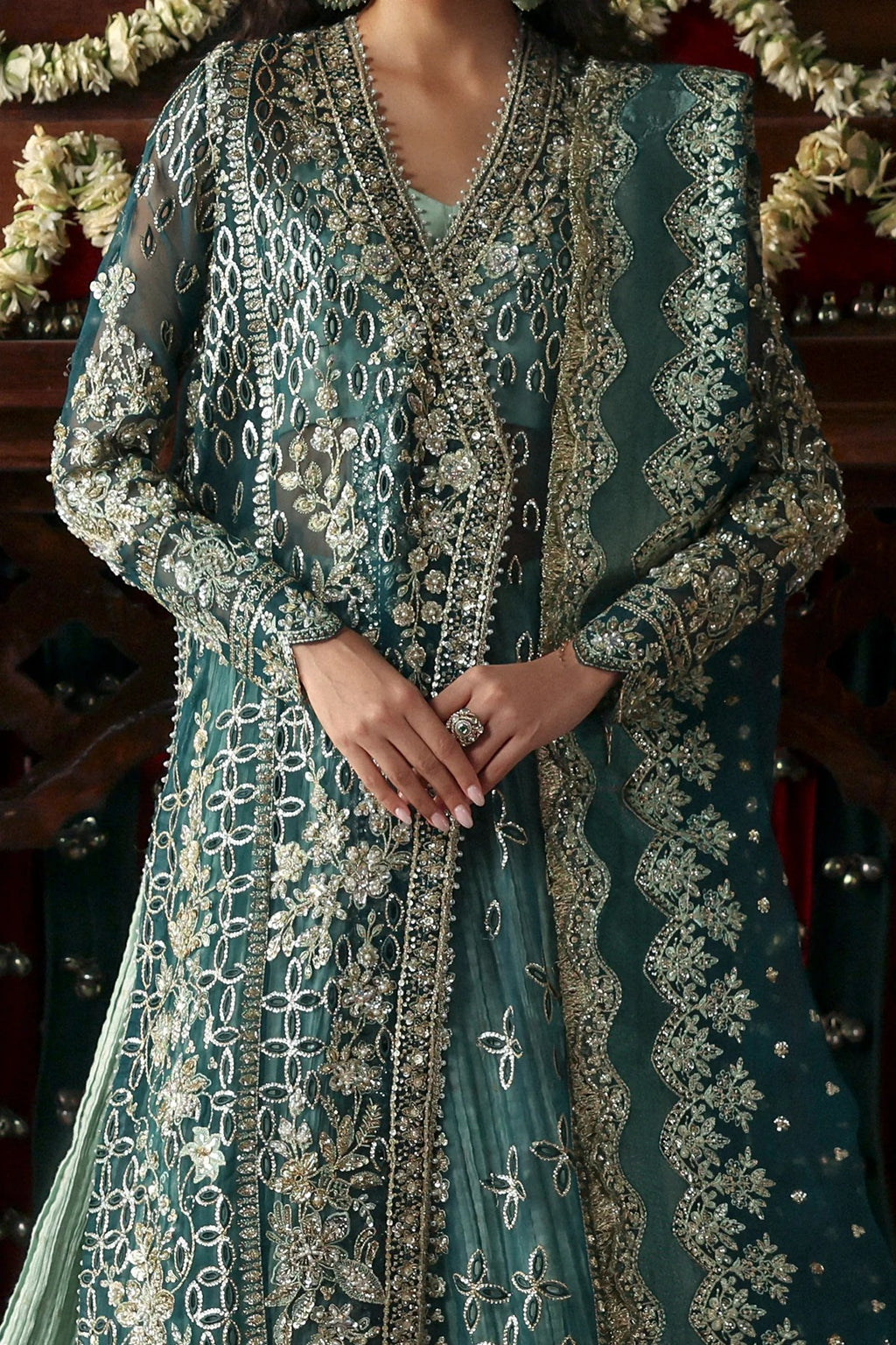 A close-up of a woman wearing an ornate teal green embroidered traditional outfit with intricate silver and gold metallic detailing, paired with a matching sheer dupatta.